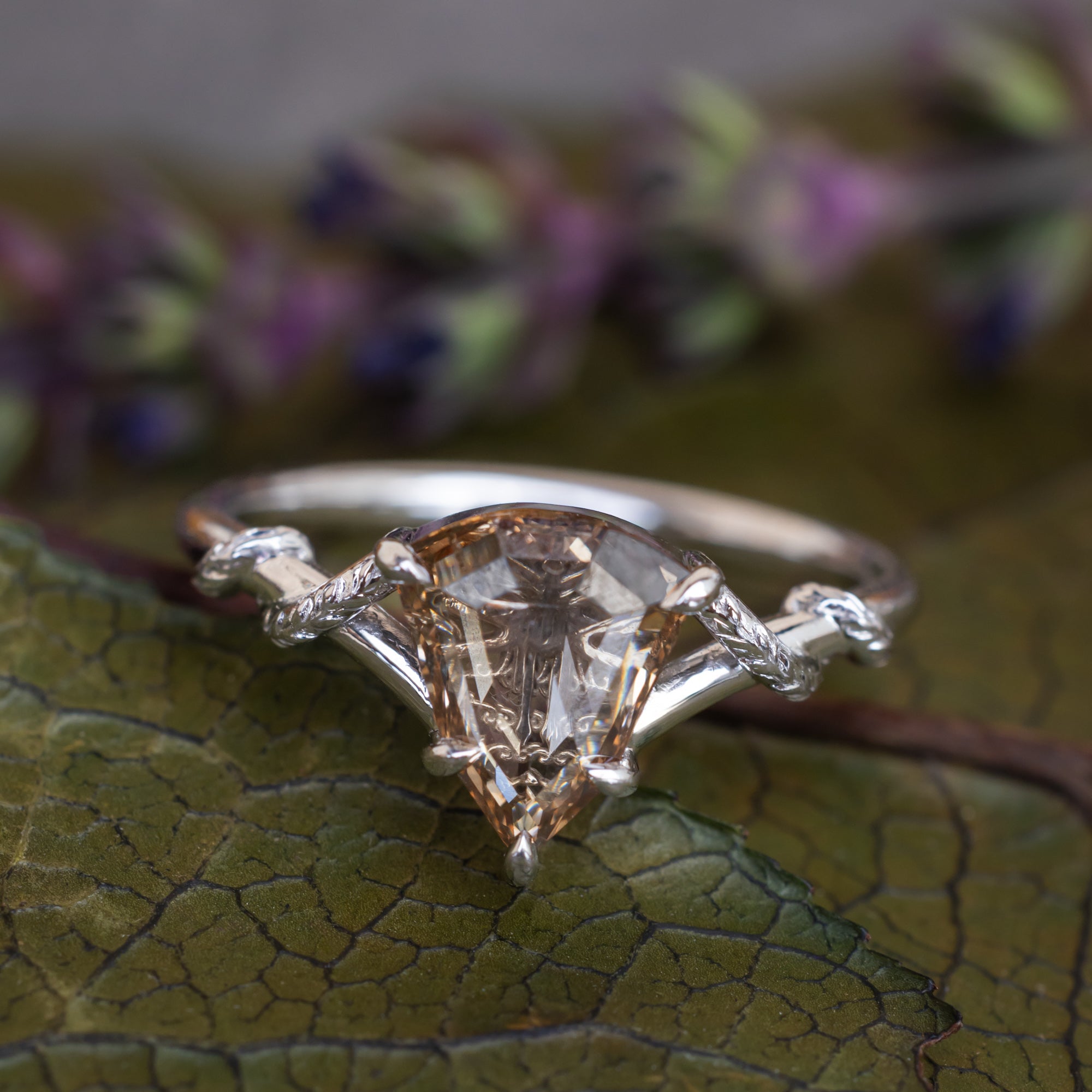 A white gold engagement ring featuring a shield-cut champagne diamond, resting on a dark green leaf with blurred purple flowers in the background