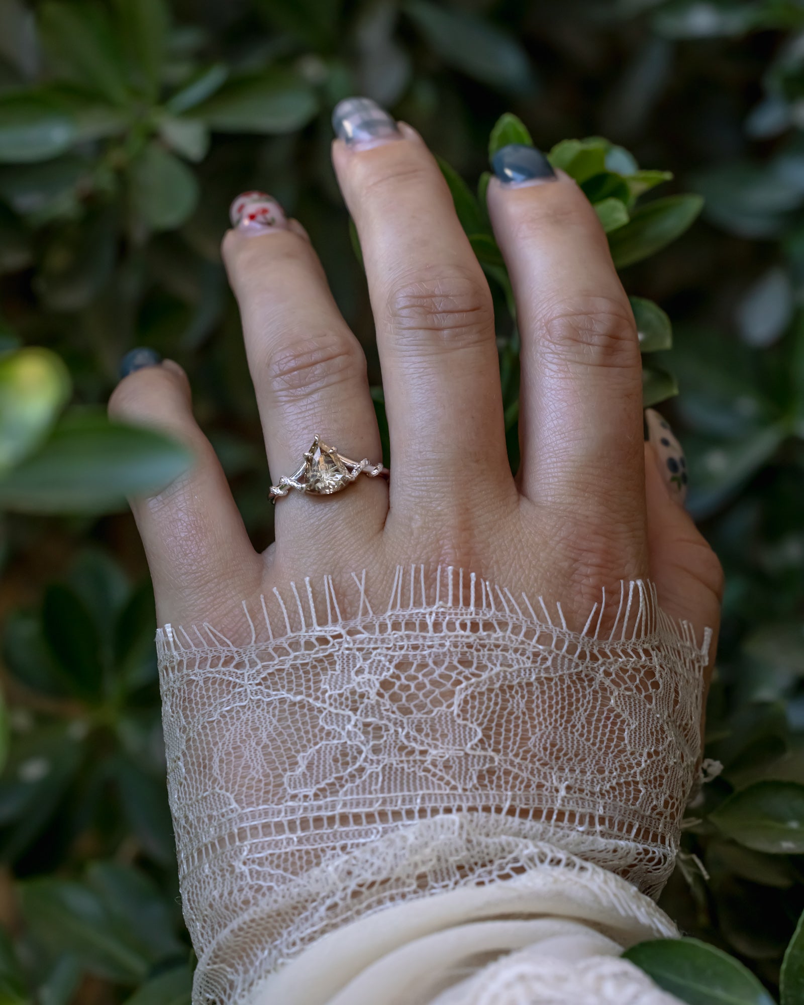 A close-up of a hand wearing a white gold ring with vine filigree, styled with a lace cuff against a background of green foliage.