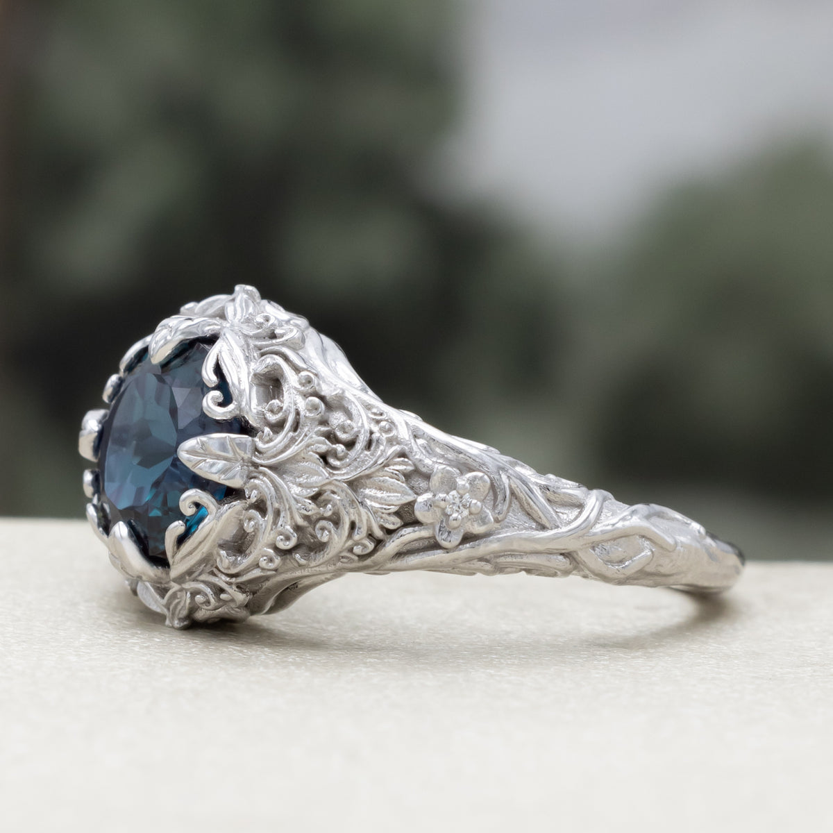 White gold ring with sculptural filigree and round alexandrite center stone against a blurred green background