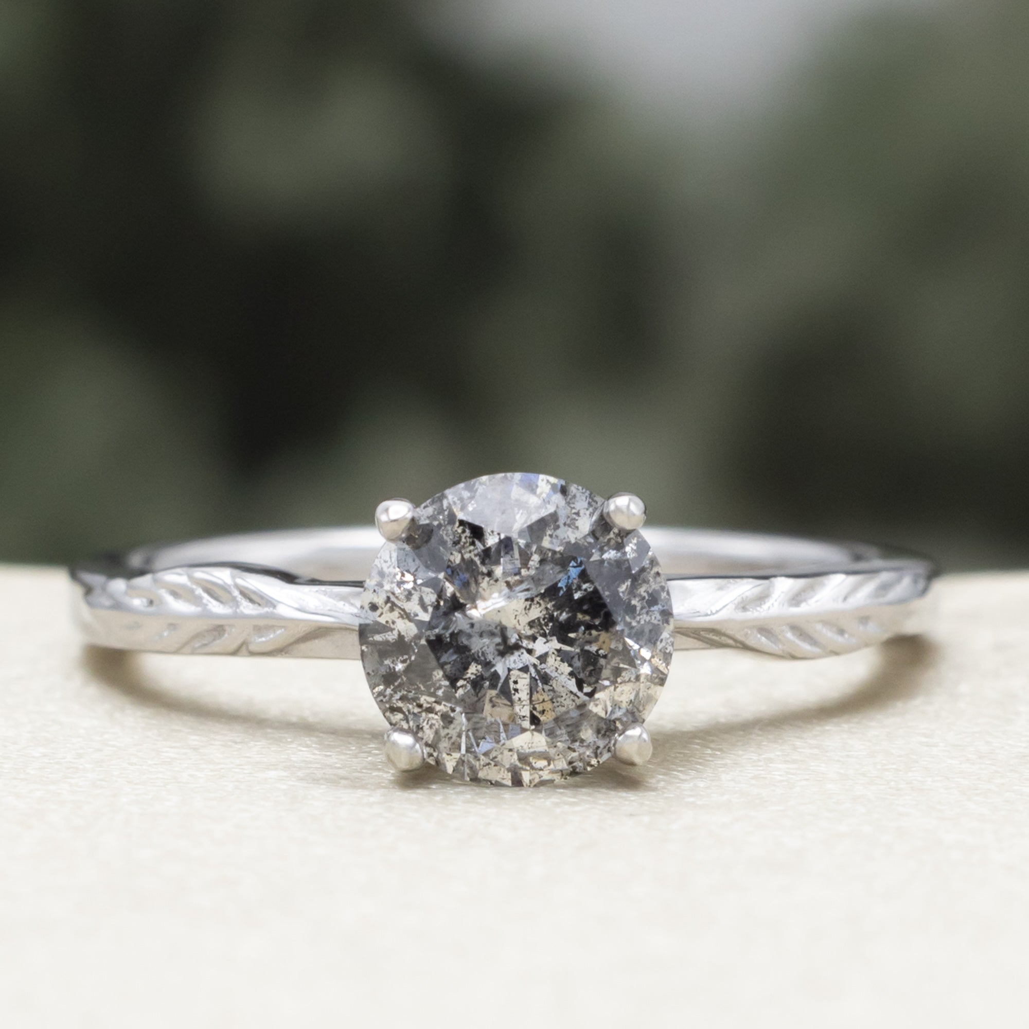White gold ring with round salt & pepper diamond in front of a blurred green background