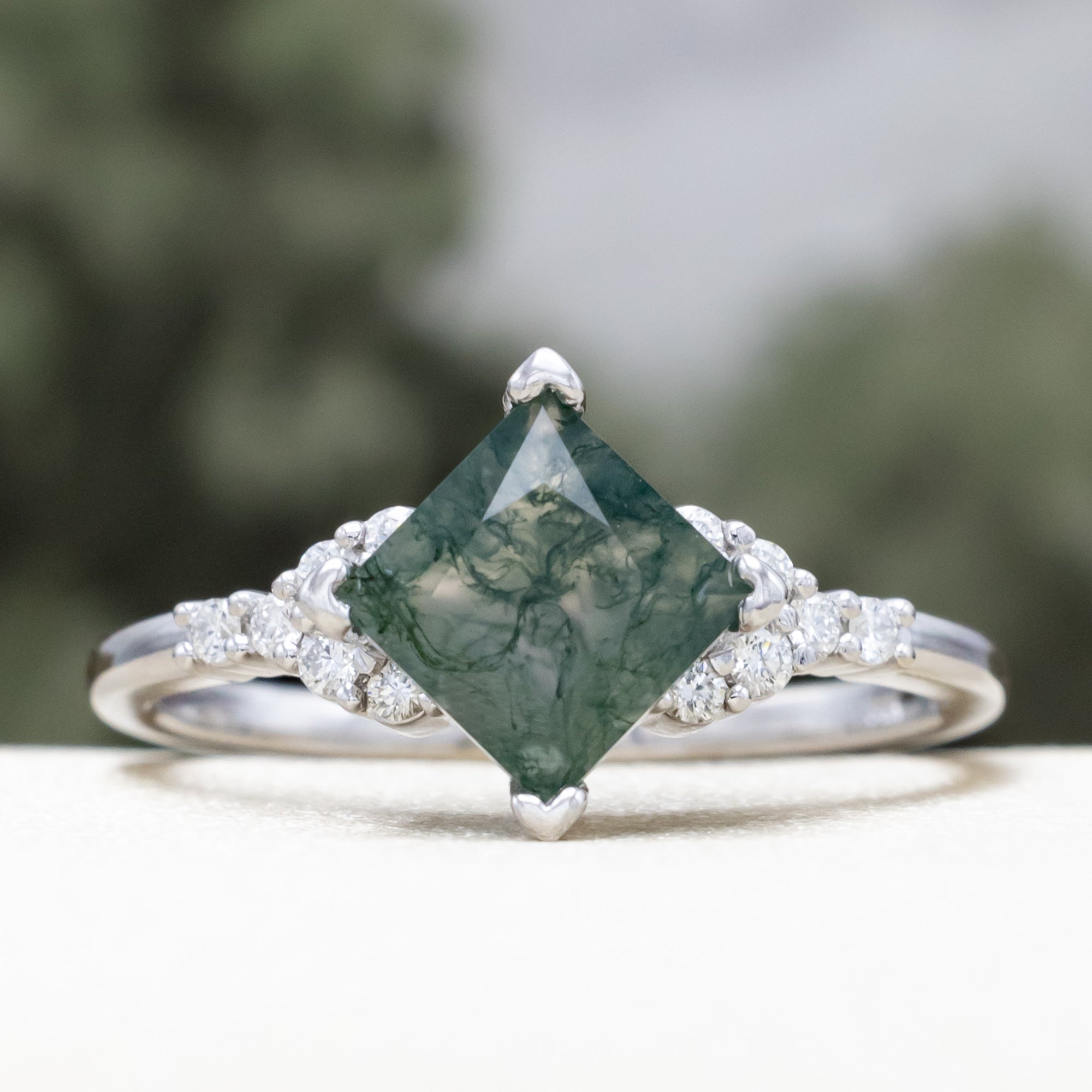 White gold ring with a moss agate center stone on a white surface with blurred greenery in the background