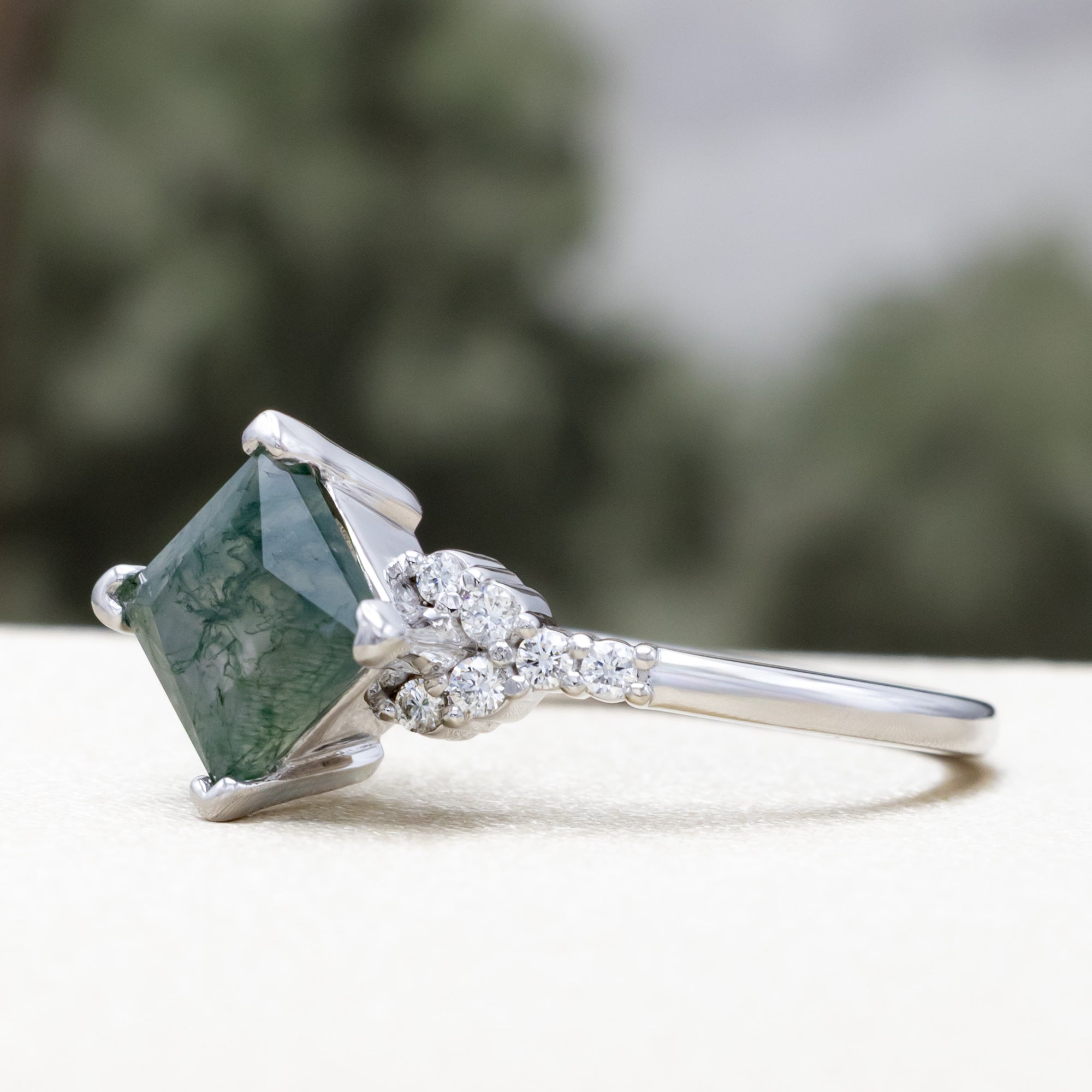 White gold ring with moss agate on a white surface with blurred greenery in the background