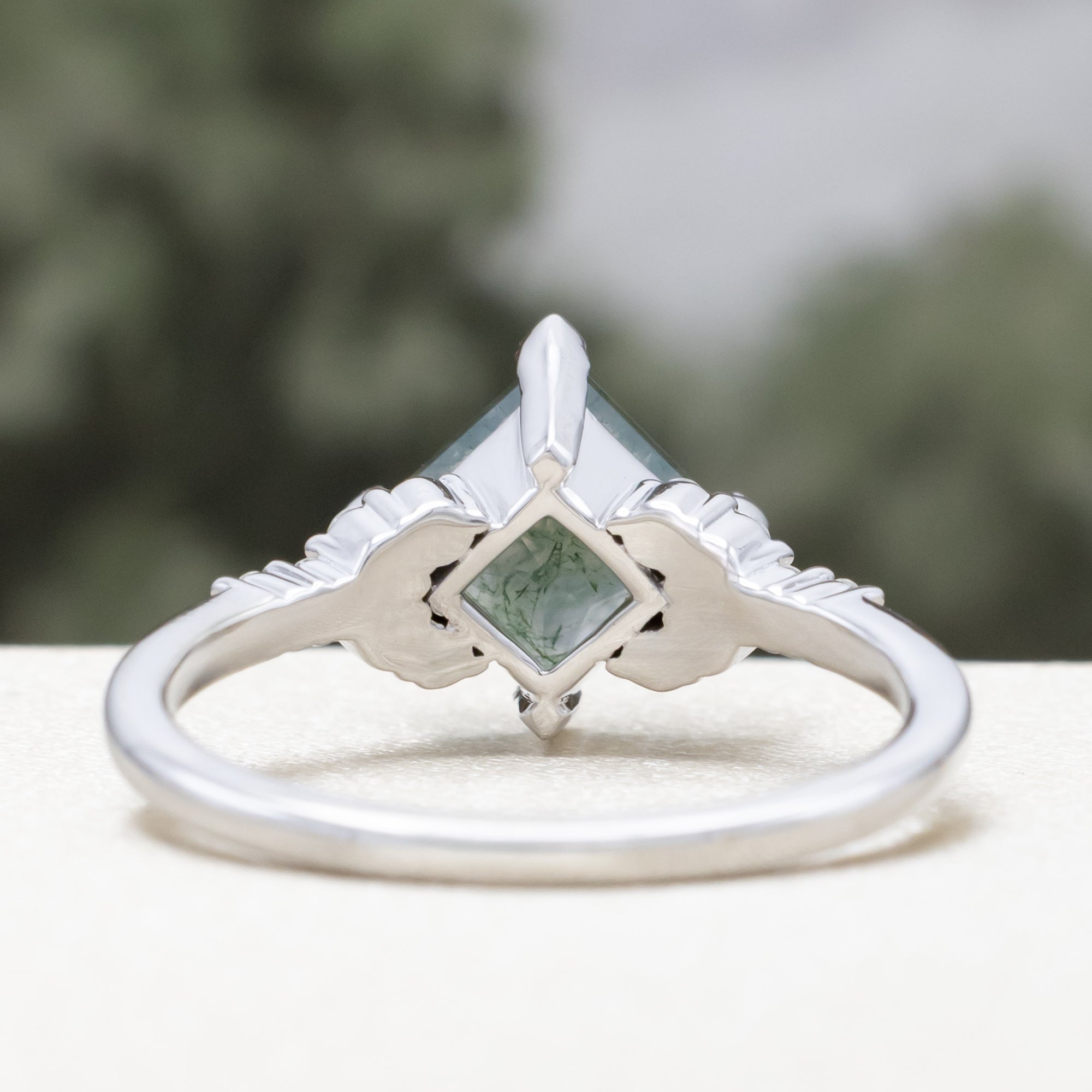 White gold ring with moss agate on a white surface with blurred greenery in the background