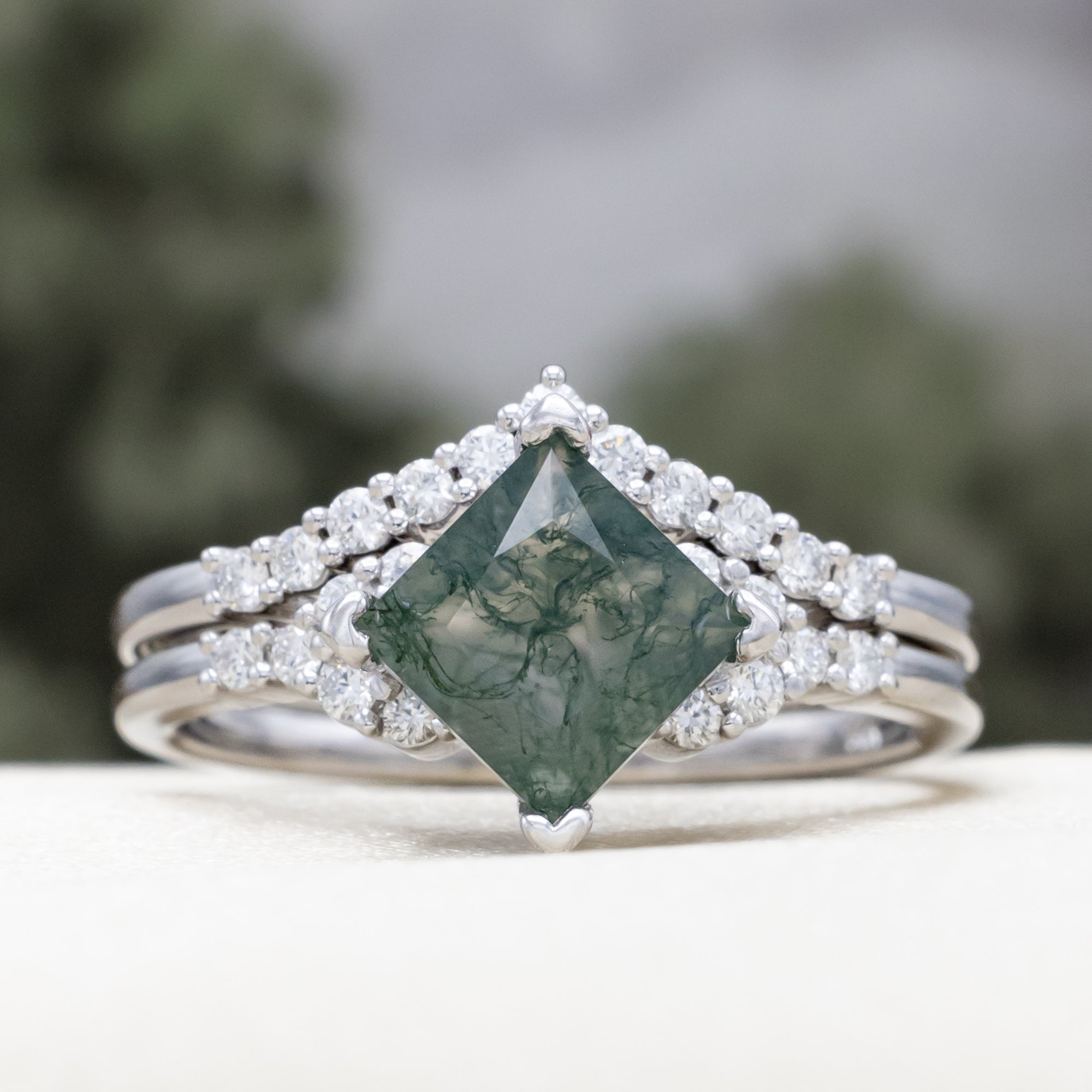 White gold ring with a moss agate center stone on a white surface with blurred greenery in the background