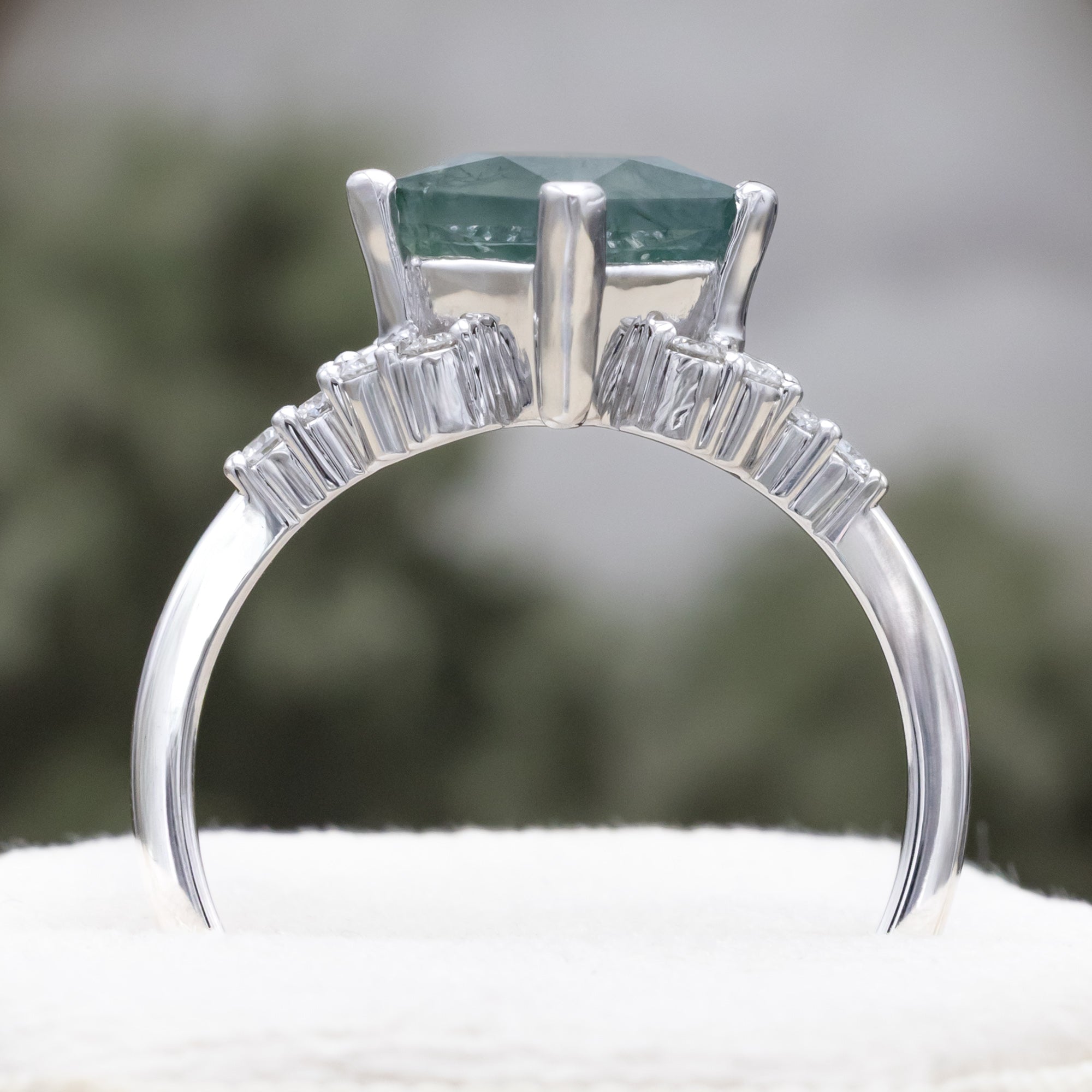 White gold ring with moss agate on a white surface with blurred greenery in the background