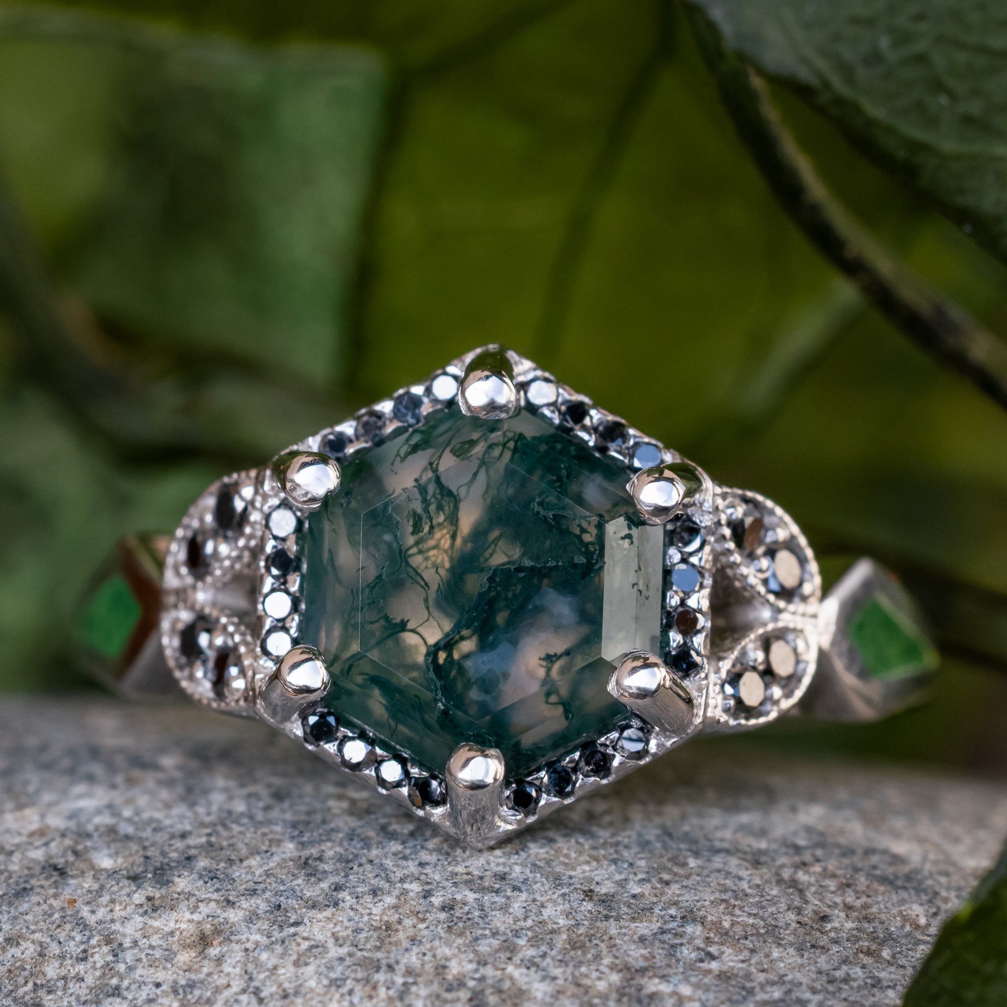 White gold ring with a moss agate center stone and black diamond accents on a stone surface with green leaves in the background