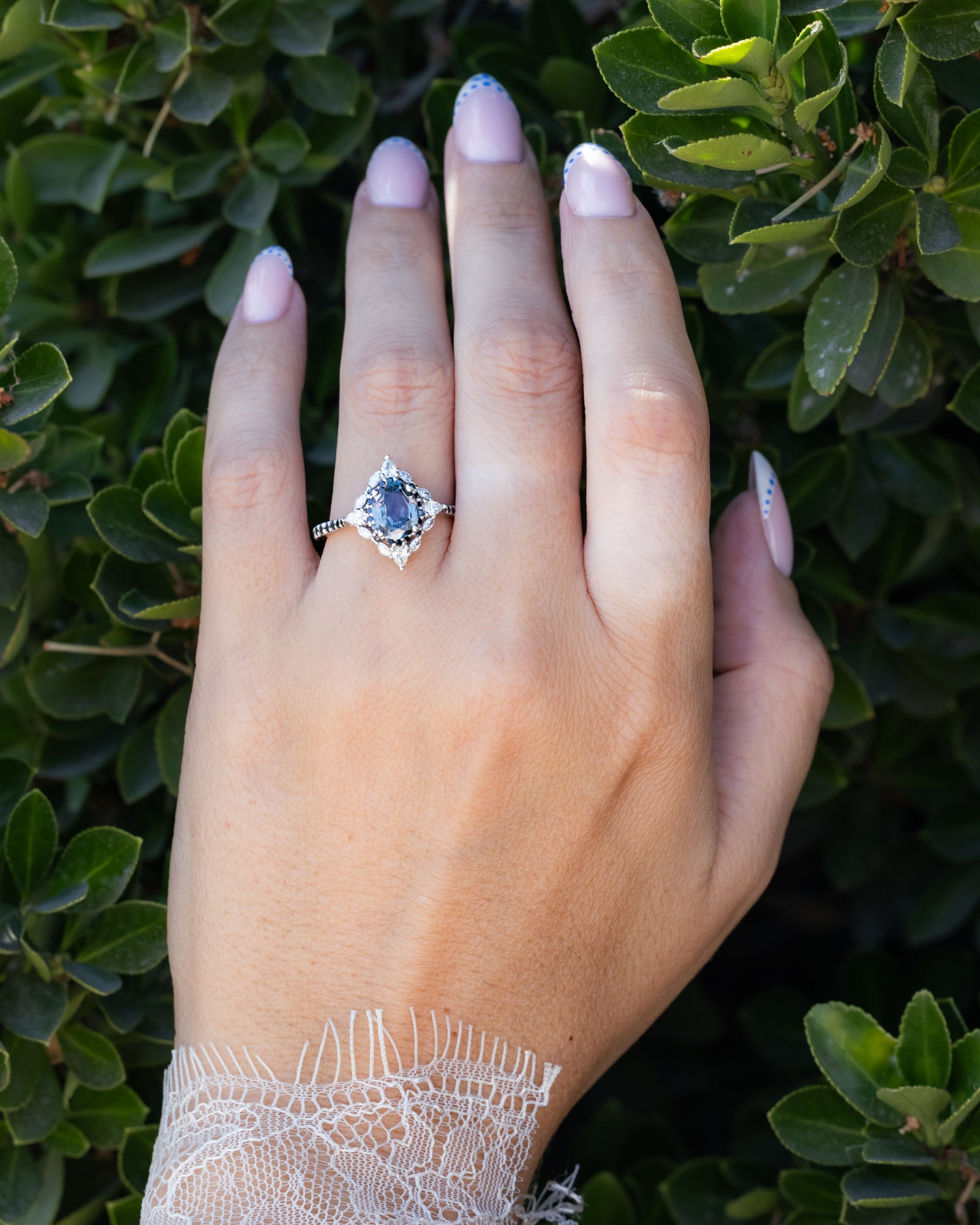 Hand wearing a ring with sapphire and diamond accents against a green leafy background