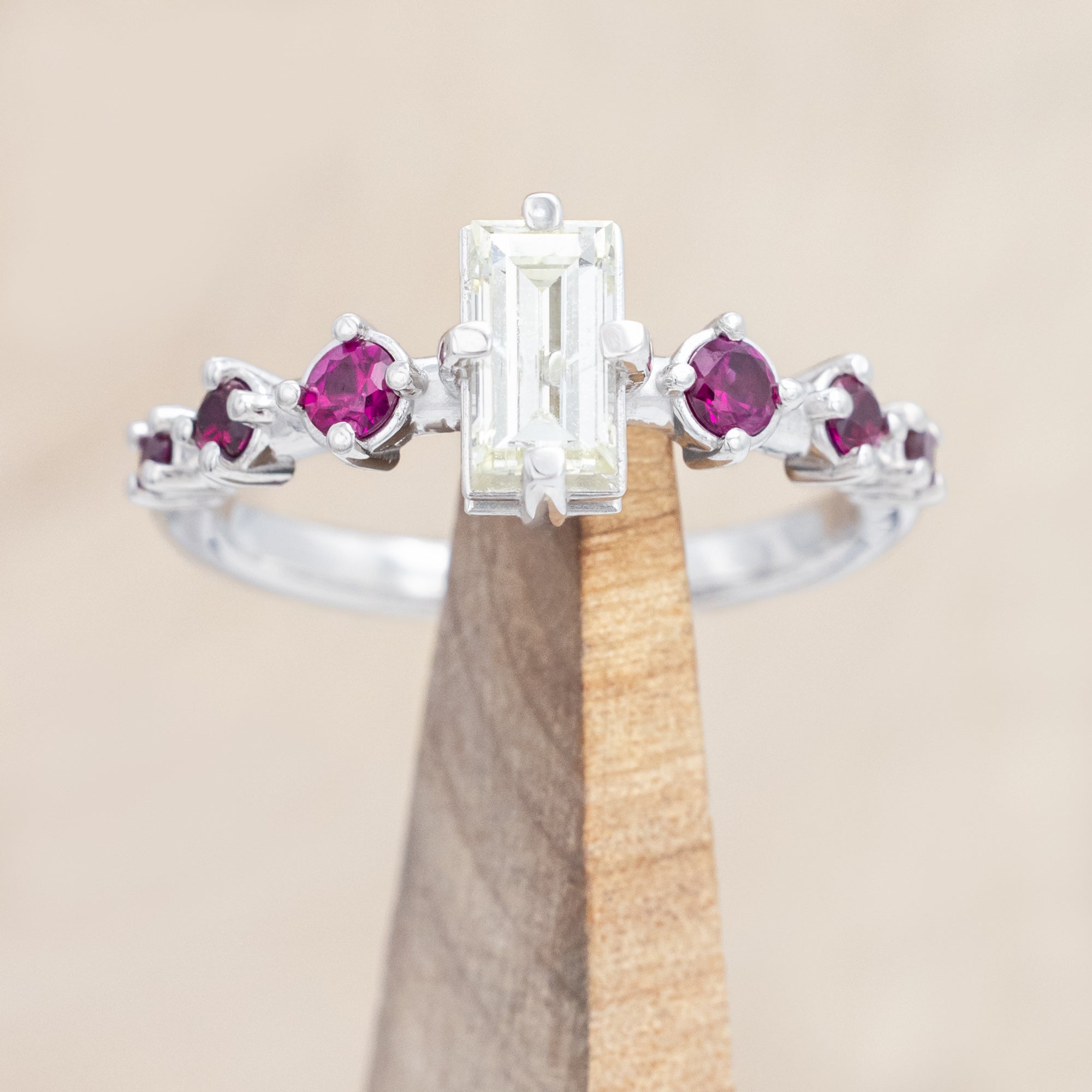14K white gold ring with central emerald-cut diamond and ruby accents on a beige background
