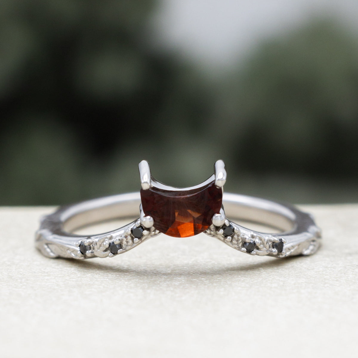 White gold crescent moon garnet tracer with vine band and black diamond accents against a blurred green background