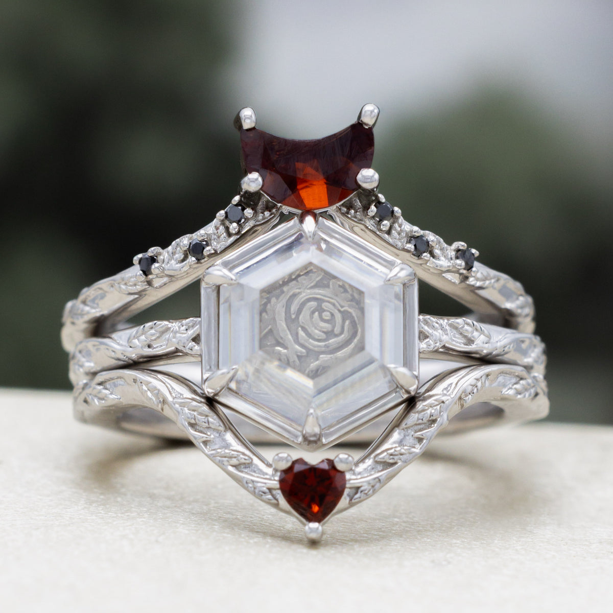 White gold engagement ring with portrait cut moissanite featuring rose design and matching garnet tracers on vine band against a blurred green background