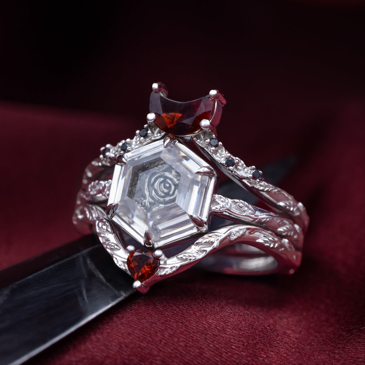 White gold engagement ring with portrait cut moissanite featuring rose design and matching garnet tracers on vine band sitting on dark red cloth