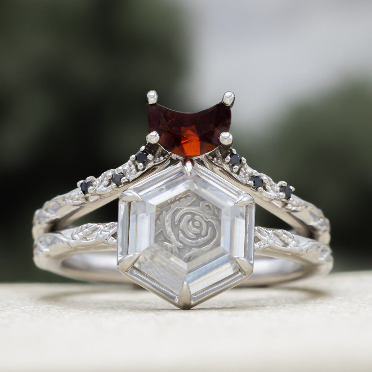 White gold engagement ring with portrait cut moissanite featuring rose design and crescent moon garnet tracer on vine band against a blurred green background