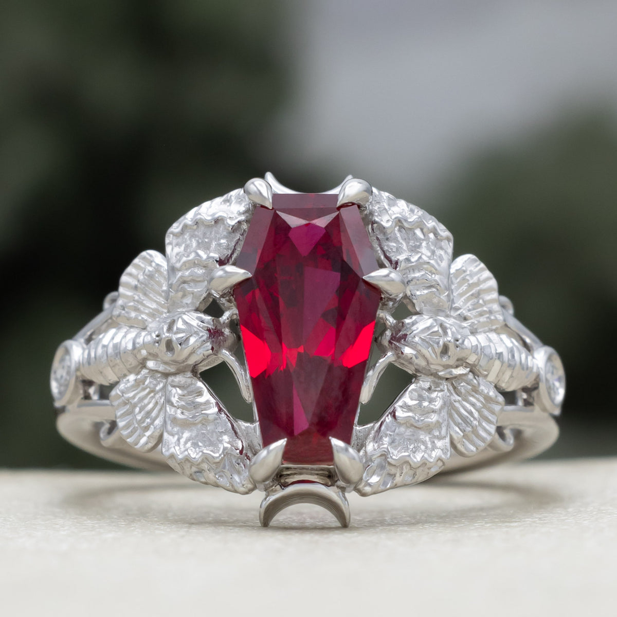 White gold engagement ring with deadhead moth filigree, coffin cut ruby center stone, and diamond accents against a blurred green background