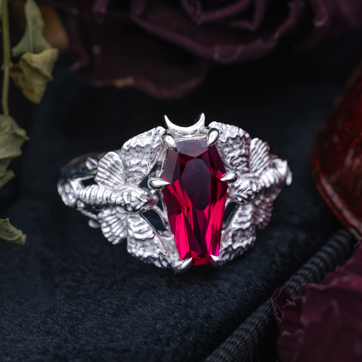 White gold engagement ring with deadhead moth filigree, coffin cut ruby center stone, and diamond accents on a black velvet surface with dried flowers in the background