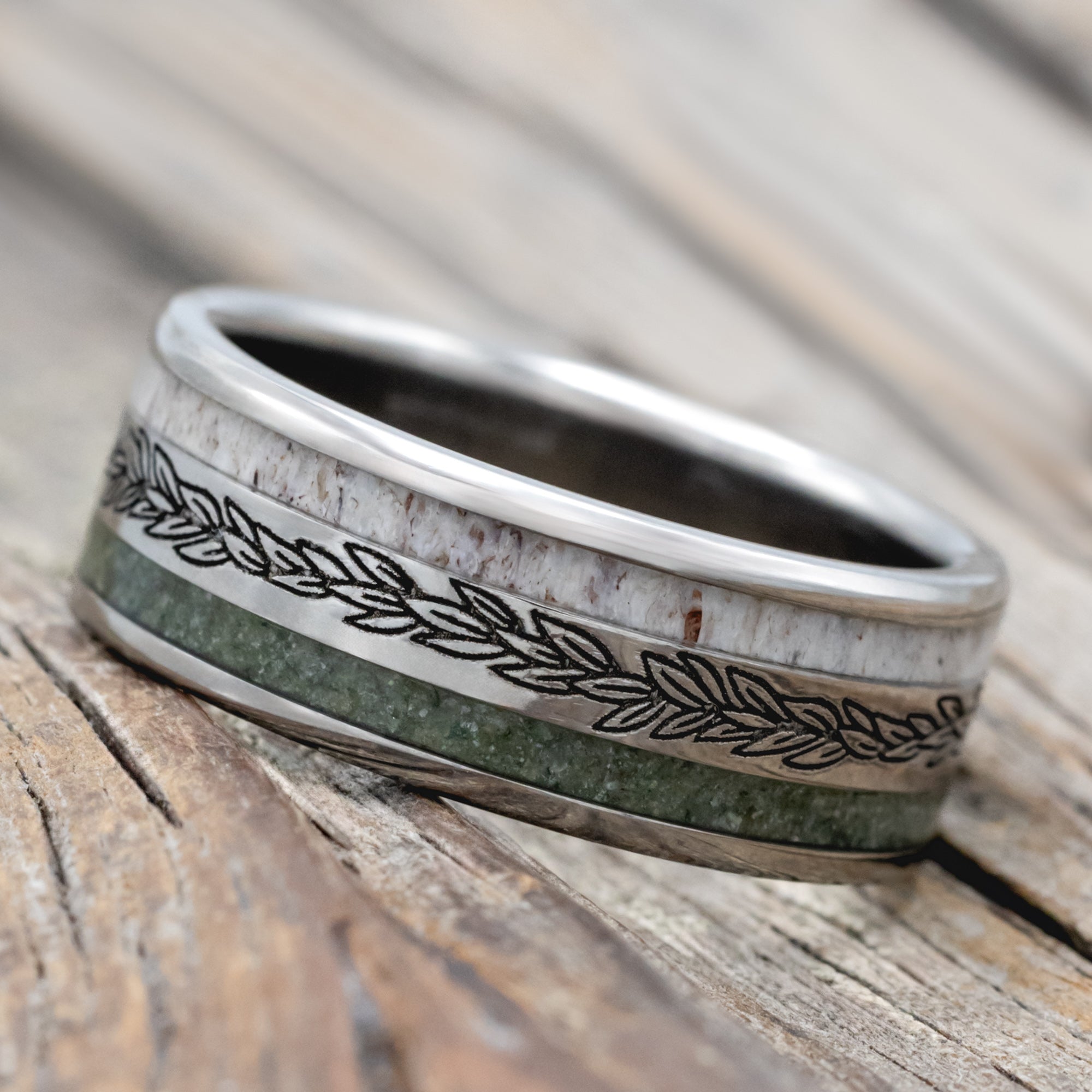 White gold ring with leaf engraving and inlays of moss agate and antler on a wooden surface
