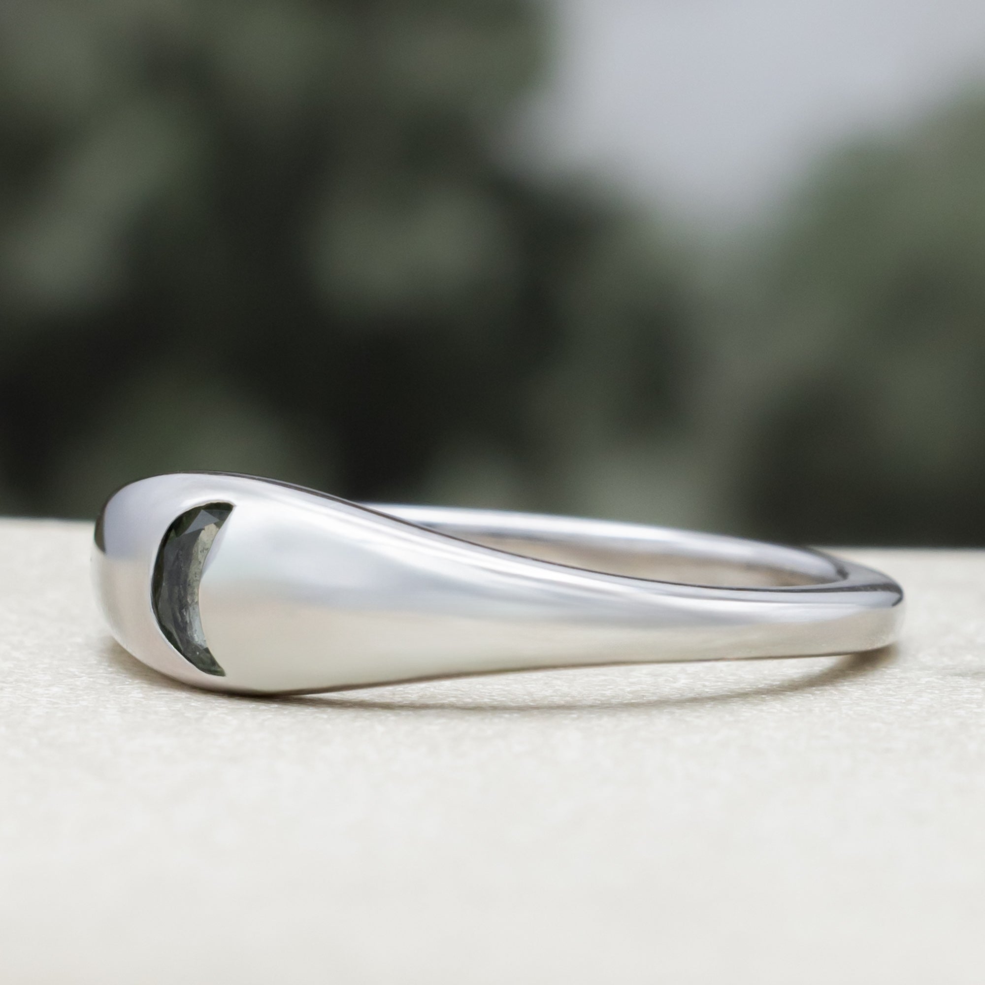 White gold ring with crescent moon shaped sapphire against a blurred green background