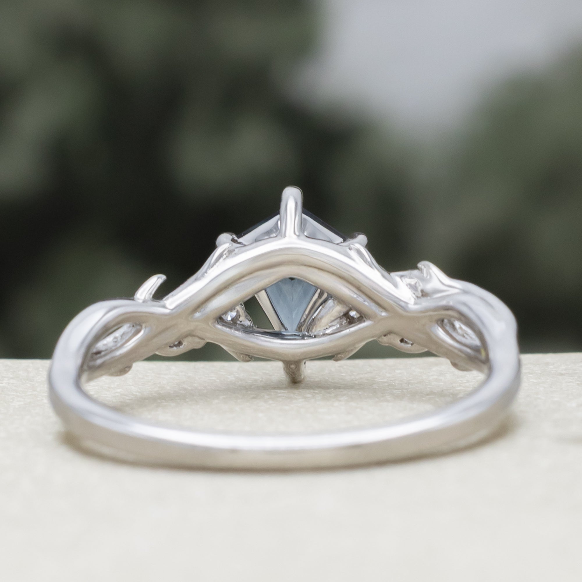 White gold ring with blue spinel center stone & diamond accents against a blurred green background