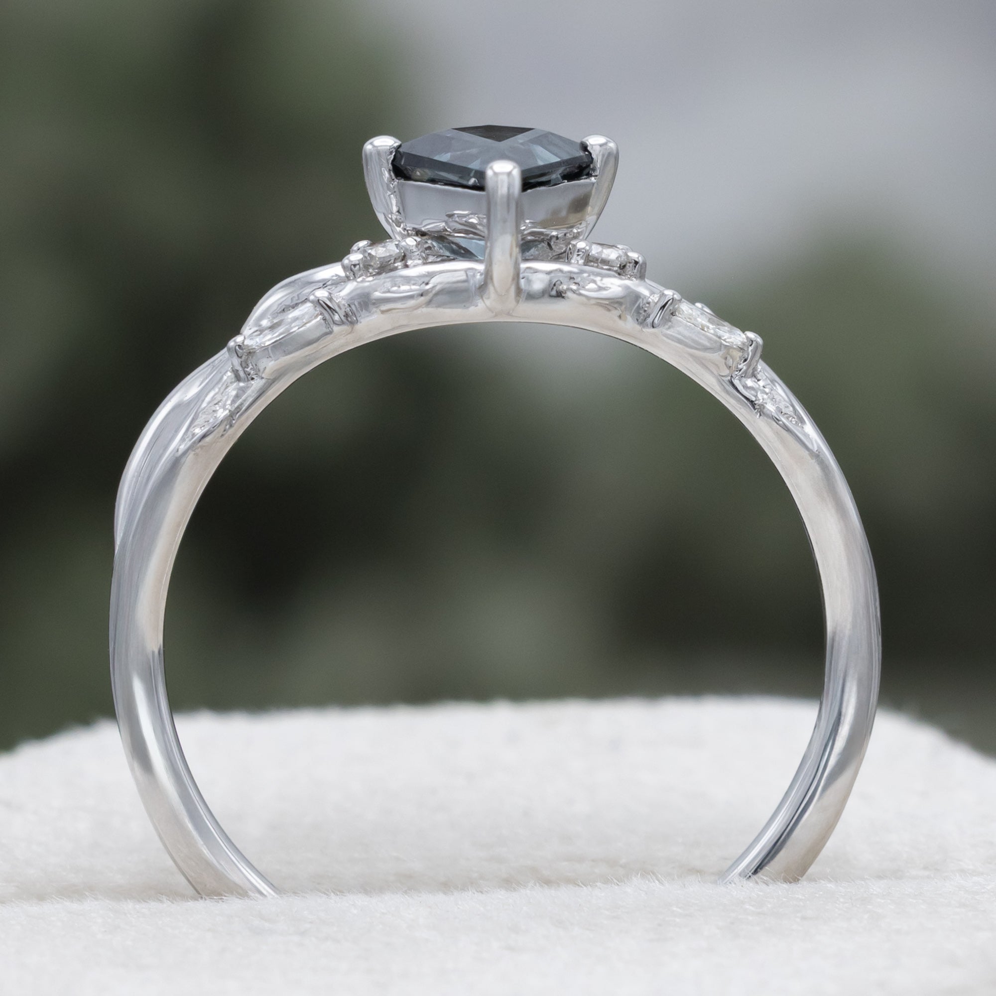 White gold ring with blue spinel center stone & diamond accents against a blurred green background