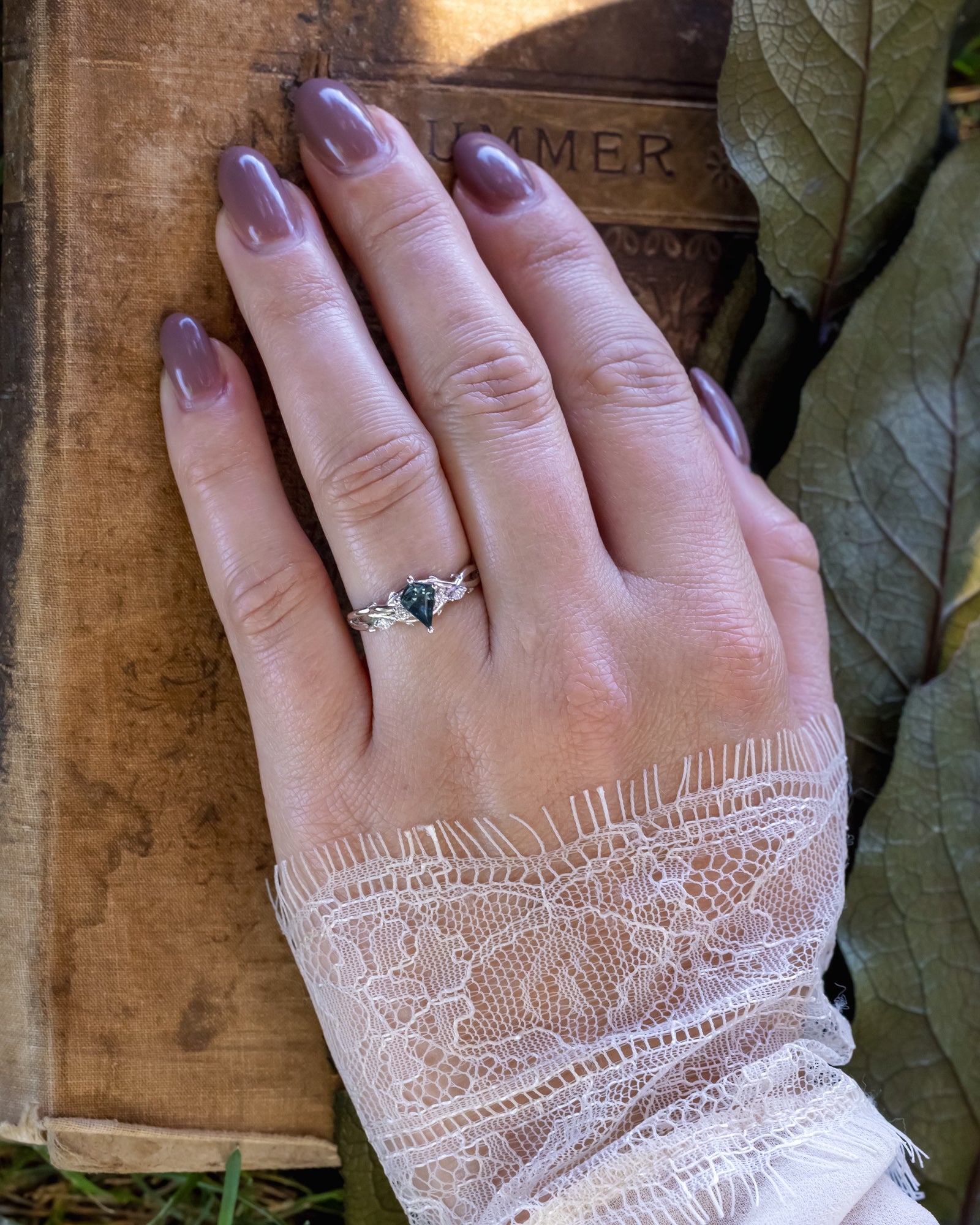 Hand wearing a ring with a blue spinel on a rustic surface with leaves in the background
