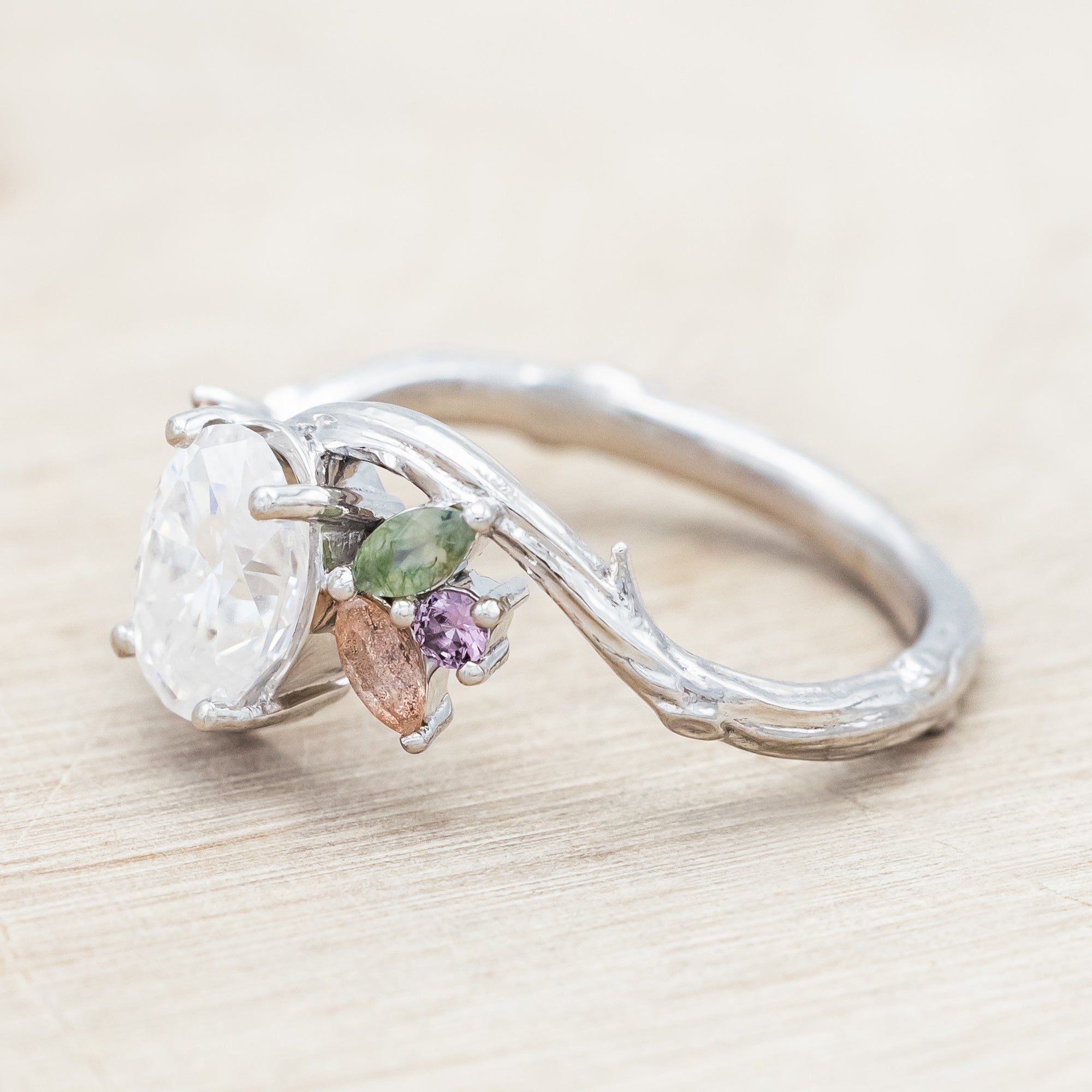 White gold ring with moissanite and smaller spinel, moss agate and sunstone accents on a wooden surface