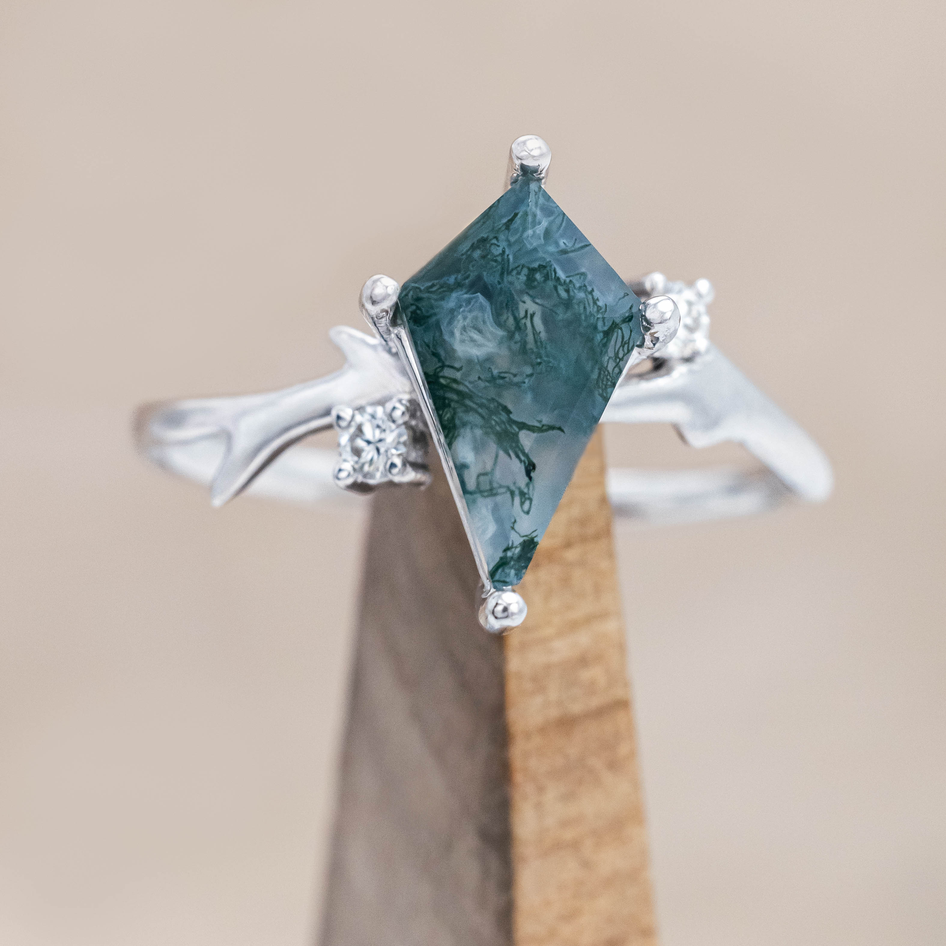 White gold ring with kite cut green moss agate on a wooden stand against a beige background 