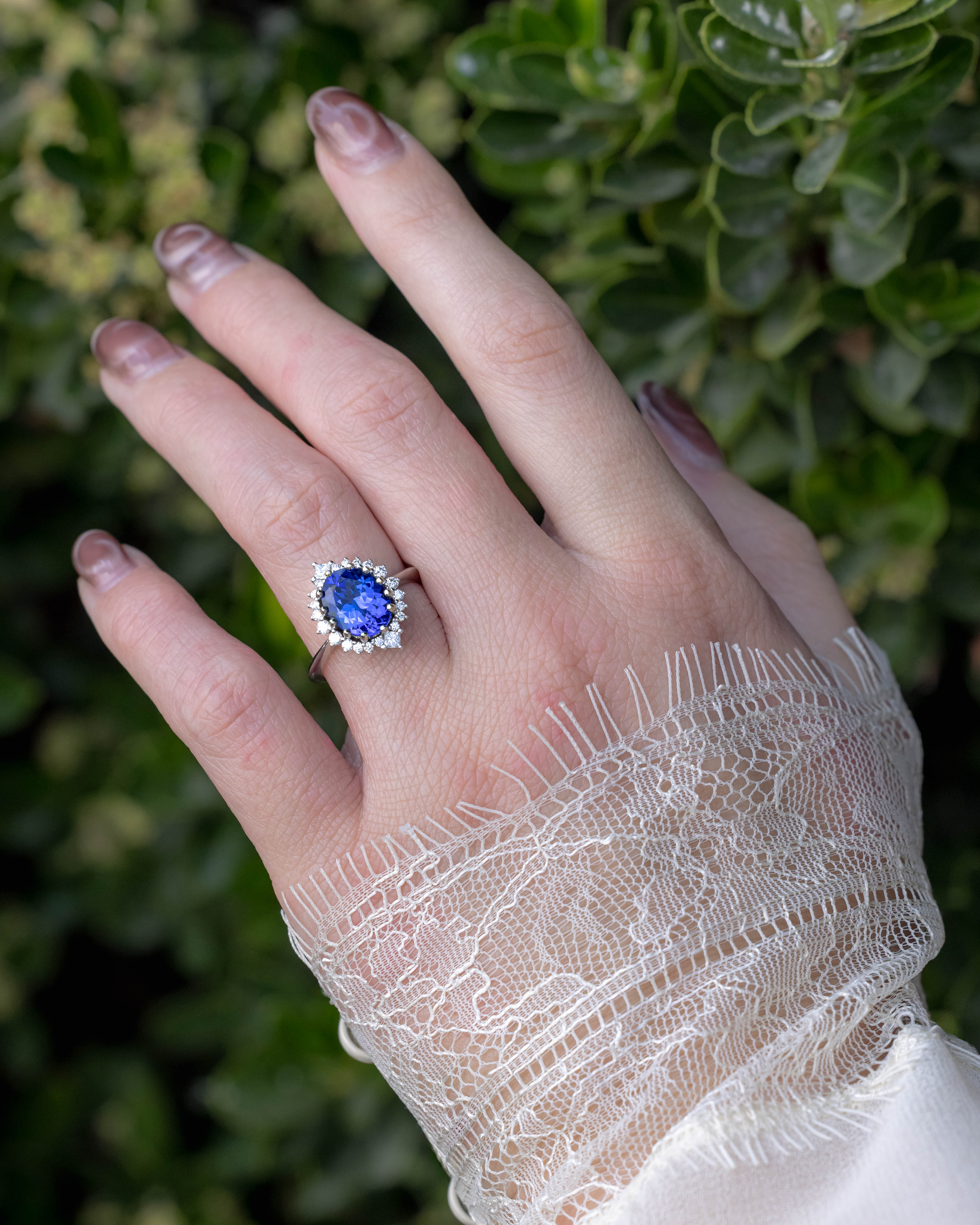 Hand wearing a ring with a blue tanzanite center stone against a green leafy background