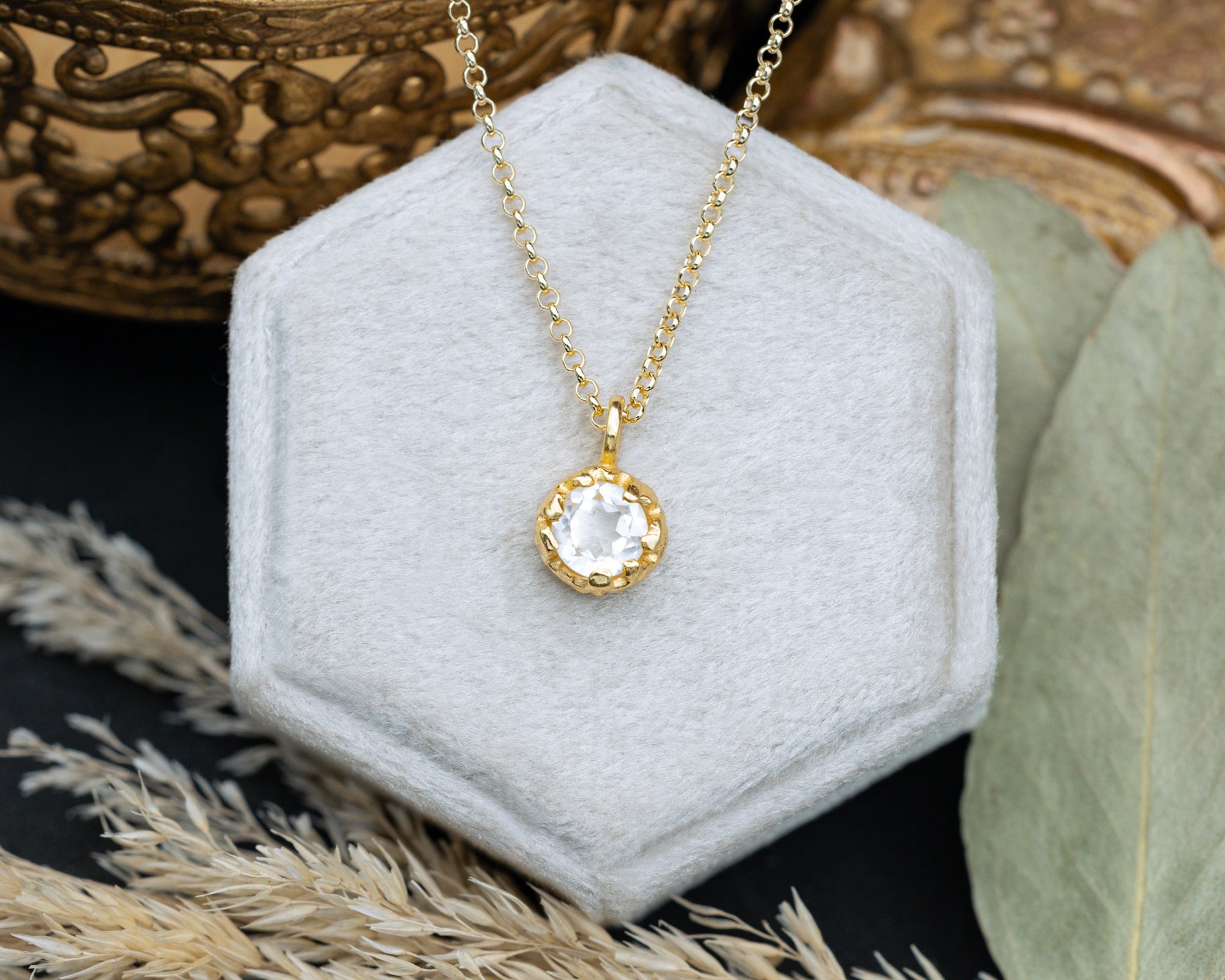 Gold necklace with a white topaz pendant on a white hexagonal stand with a decorative background