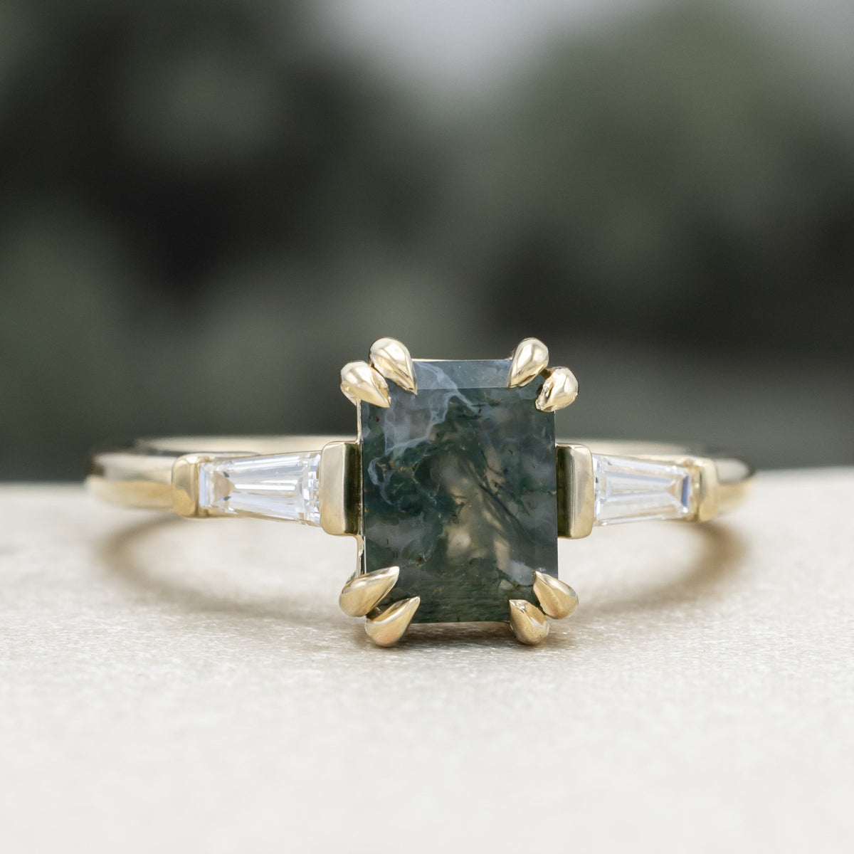 Gold ring with emerald cut moss agate center stone & diamond accents against a blurred green background