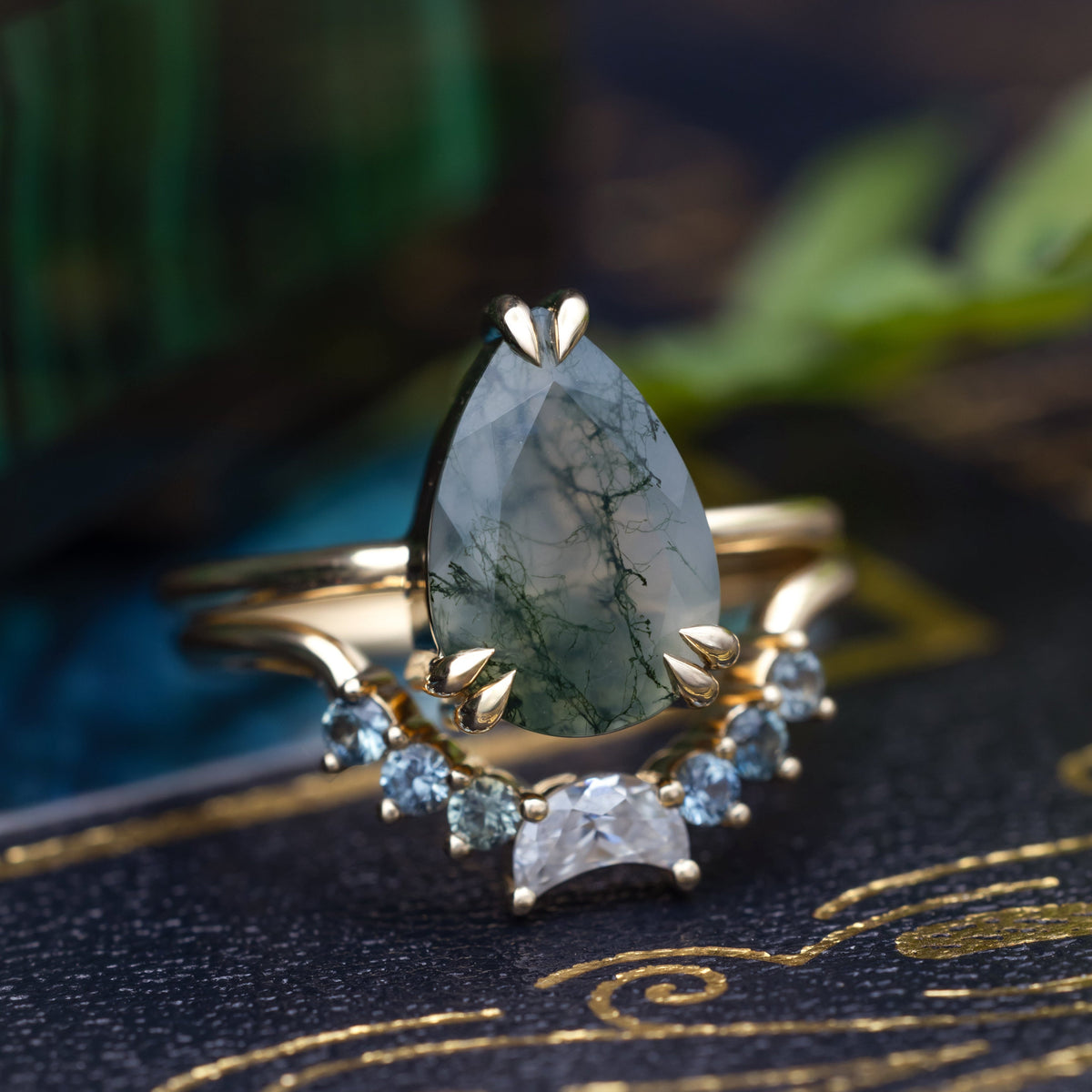 Gold moss agate engagement ring featuring tracer with moissanite and Montana sapphire accents against a moody blurred background
