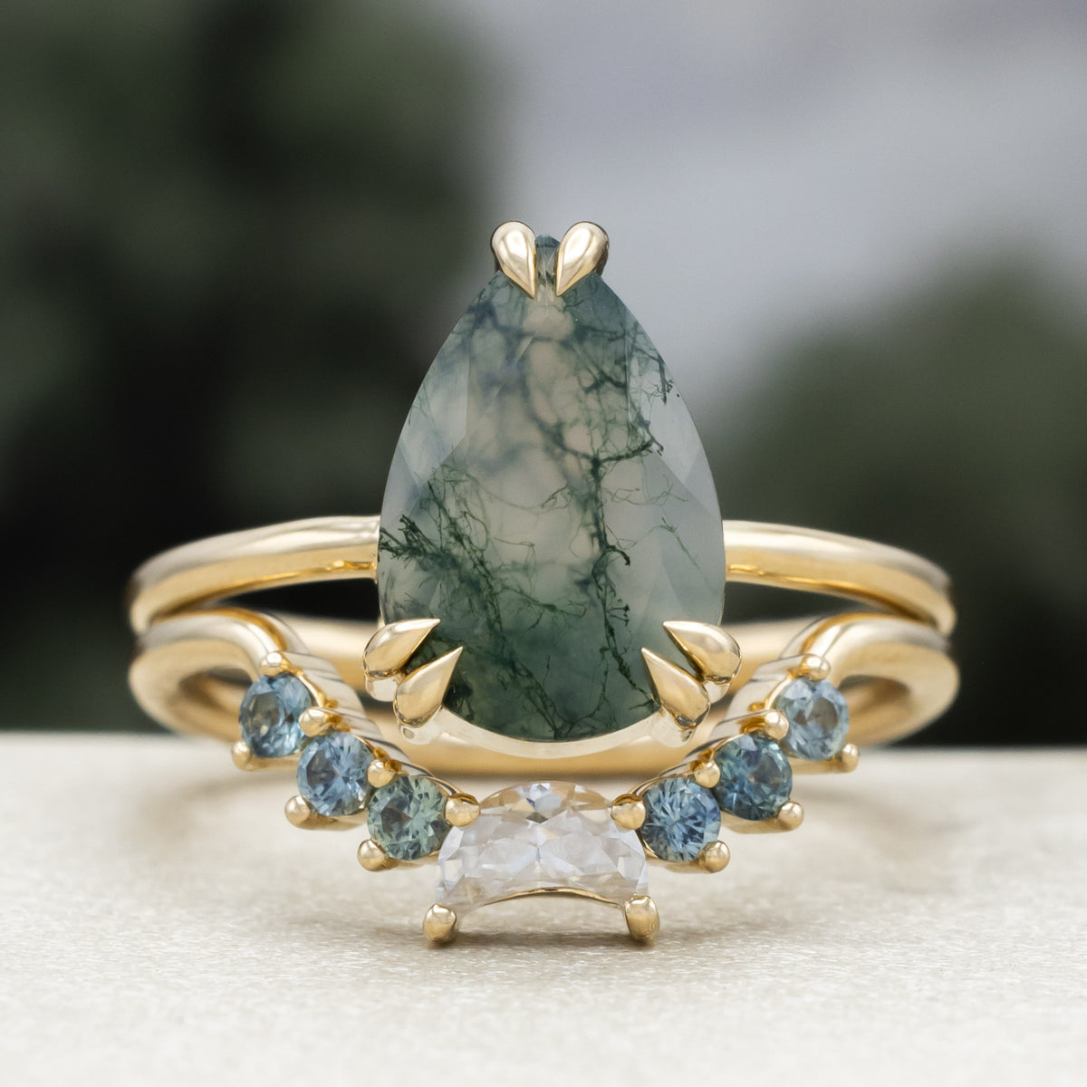 Gold ring with pear cut moss agate featuring tracer with moissanite and Montana sapphire accents against a blurred green background