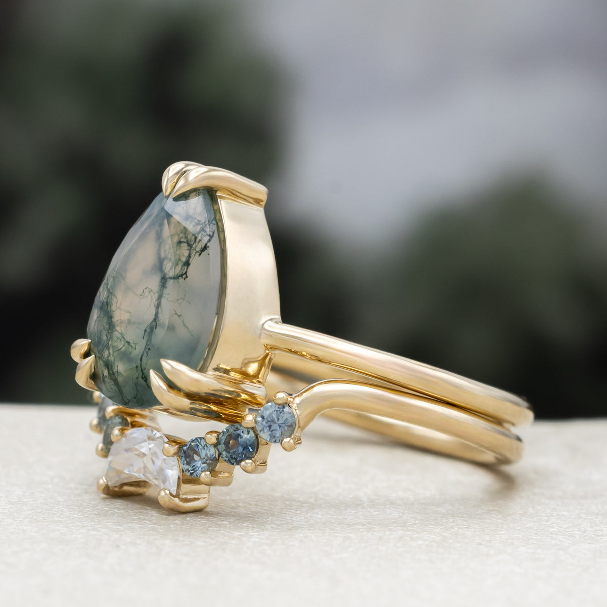 Gold ring with pear cut moss agate featuring tracer with moissanite and Montana sapphire accents against a blurred green background