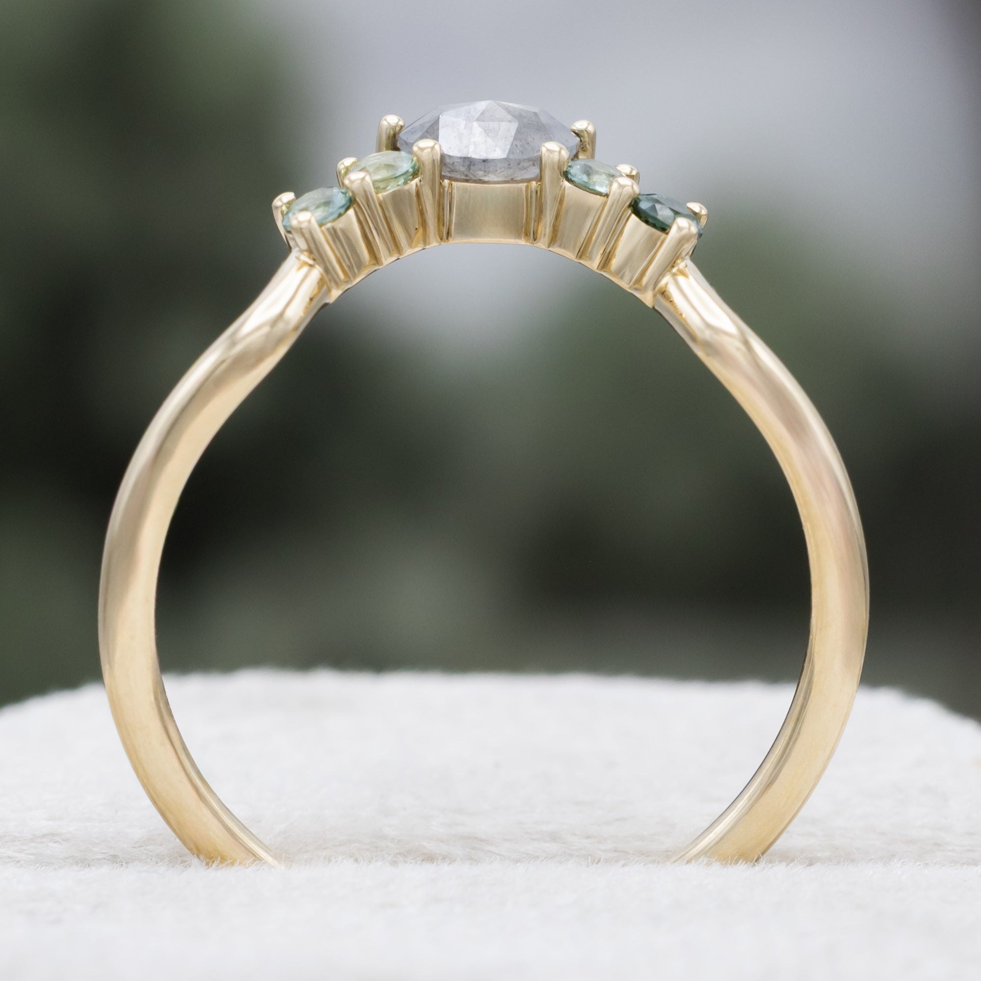 A gold ring tracer with round torumaline and crescent moon cut salt & pepper diamond on a blurred green background