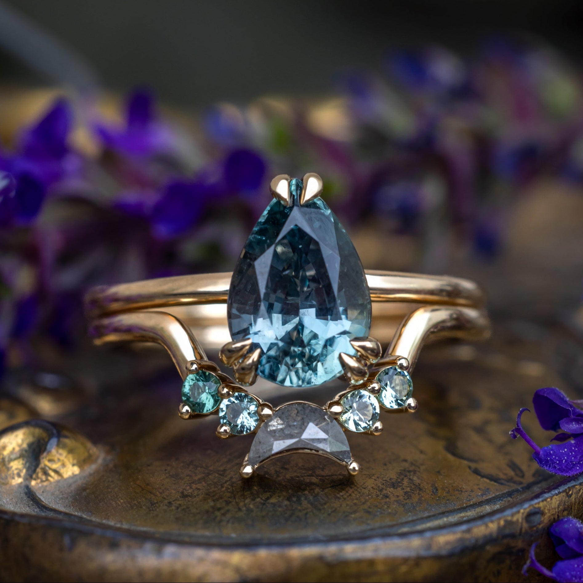 A gold ring featuring a pear-cut blue sapphire with tourmaline and salt & pepper diamond tracer on a textured bronze surface with blurred purple flowers in the background.