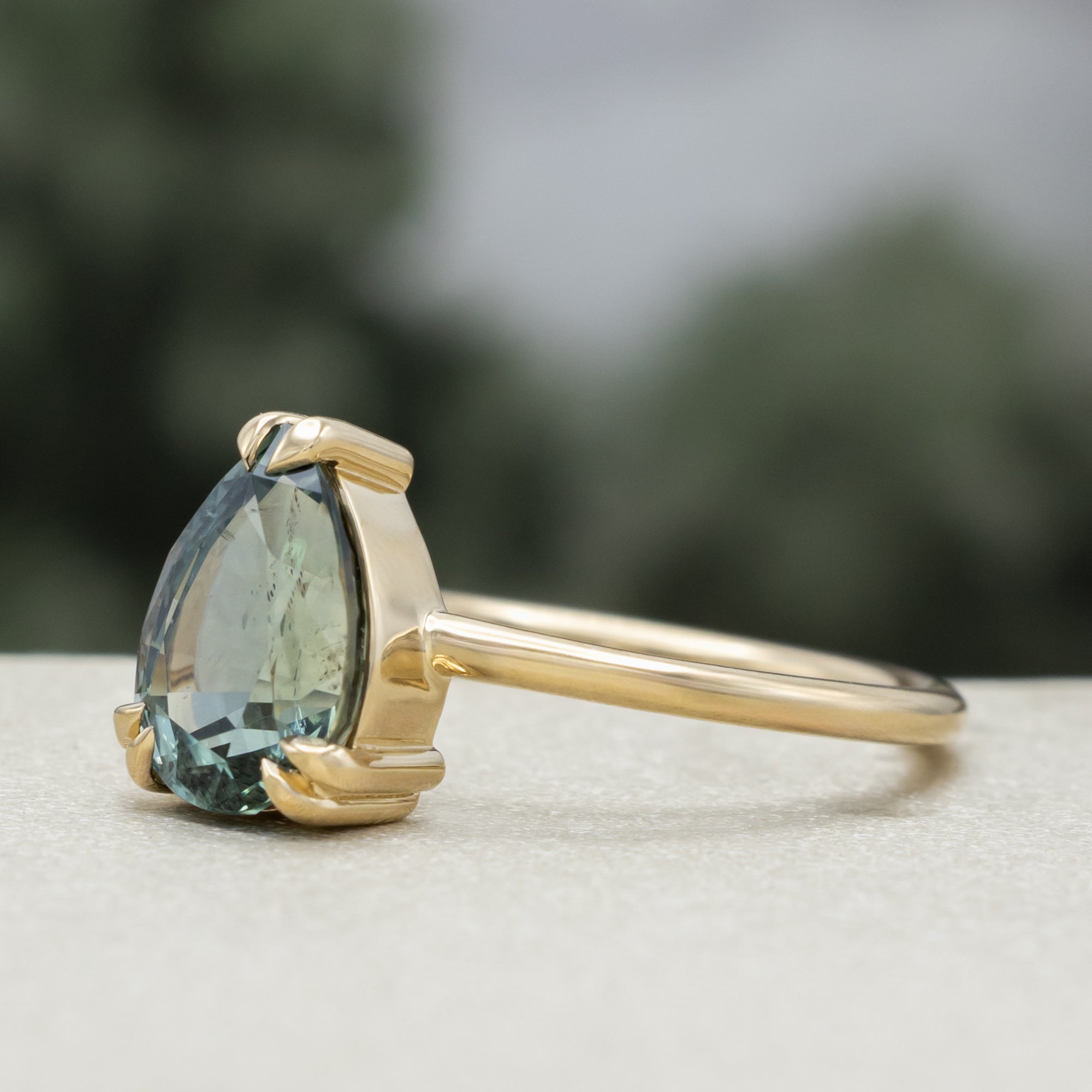 A solitaire gold ring featuring a pear-cut blue sapphire on a blurred green background.