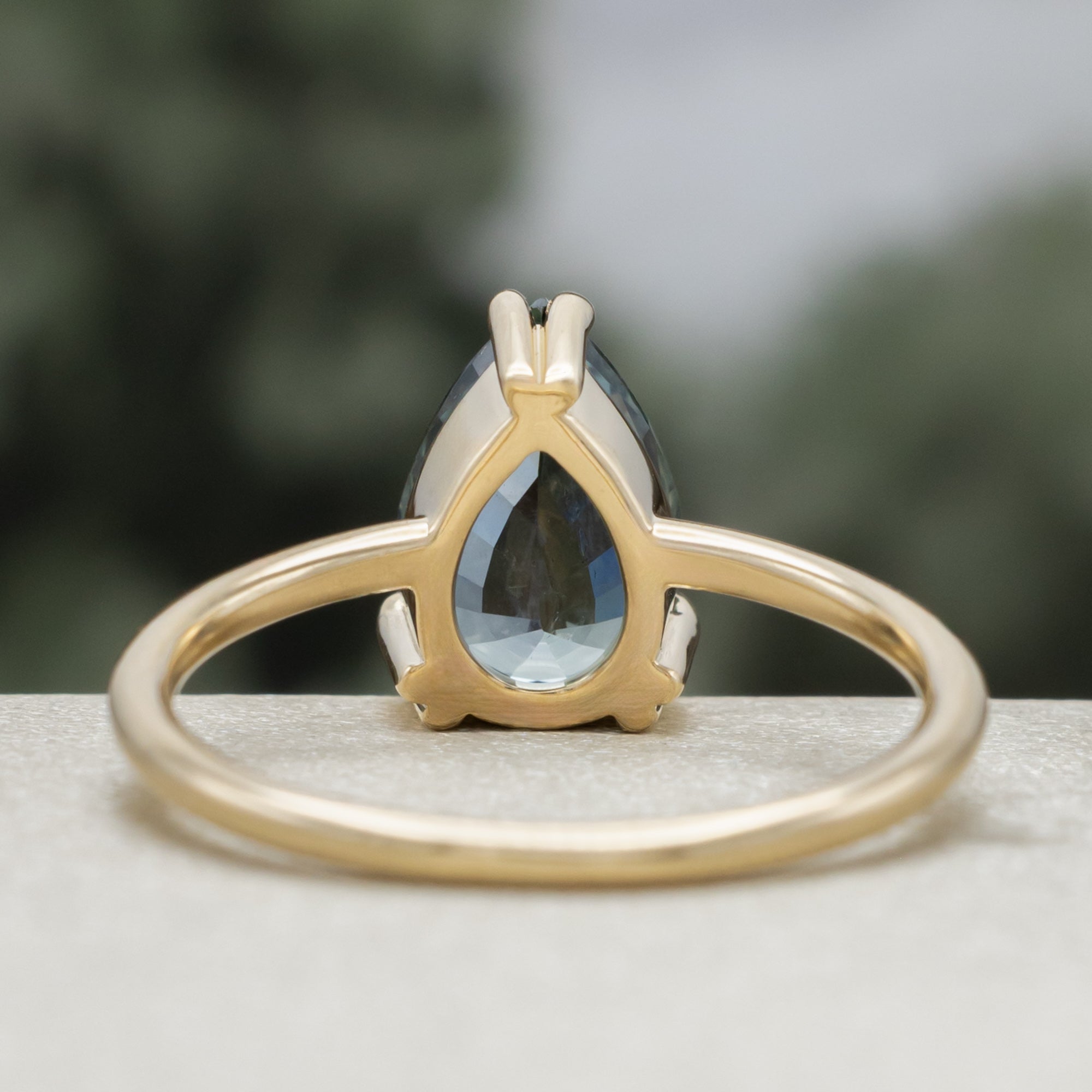 A solitaire gold ring featuring a pear-cut blue sapphire on a blurred green background.