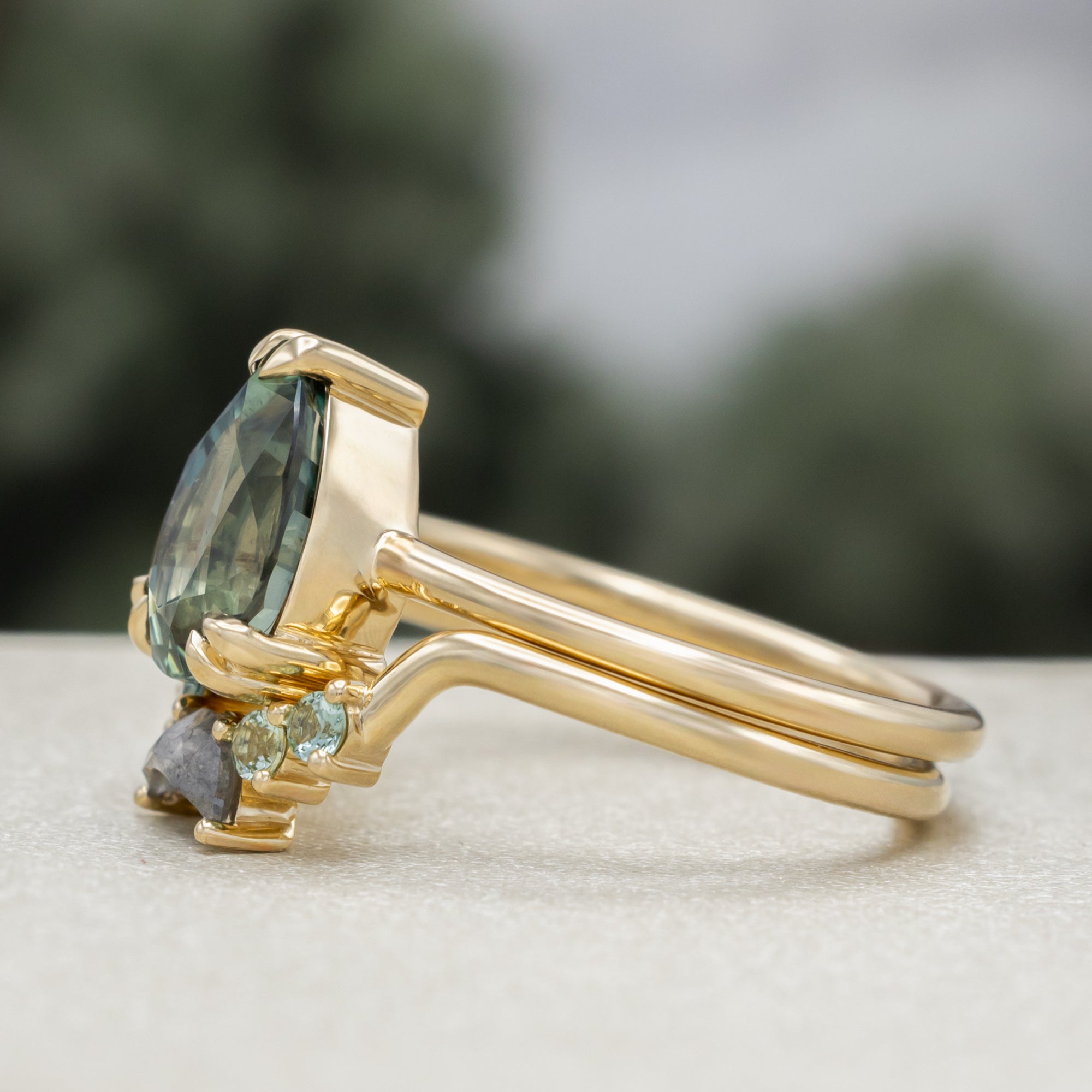 A gold ring featuring a pear-cut blue sapphire with tourmaline and salt & pepper diamond tracer on a blurred green background.