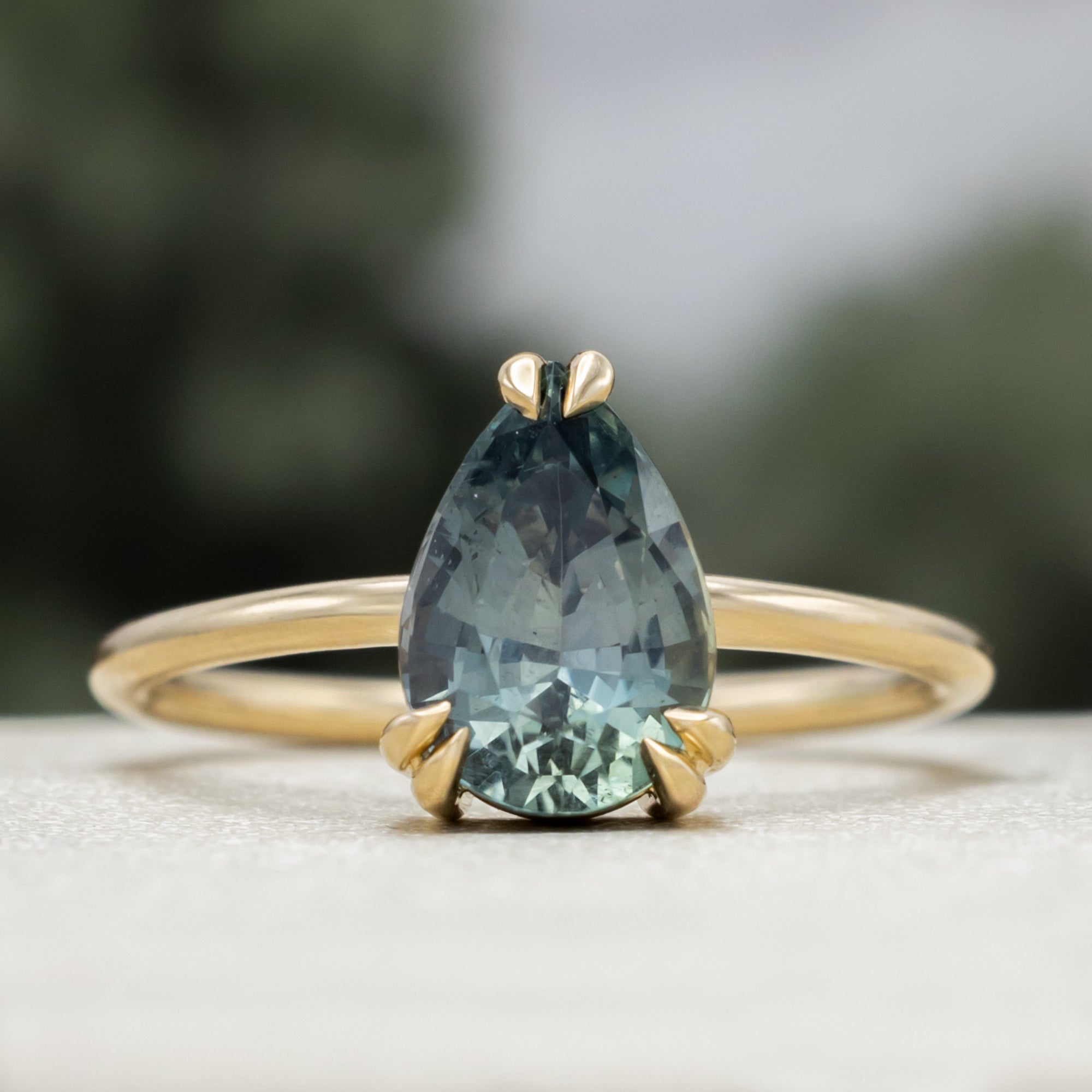 A solitaire gold ring featuring a pear-cut blue sapphire on a blurred green background.