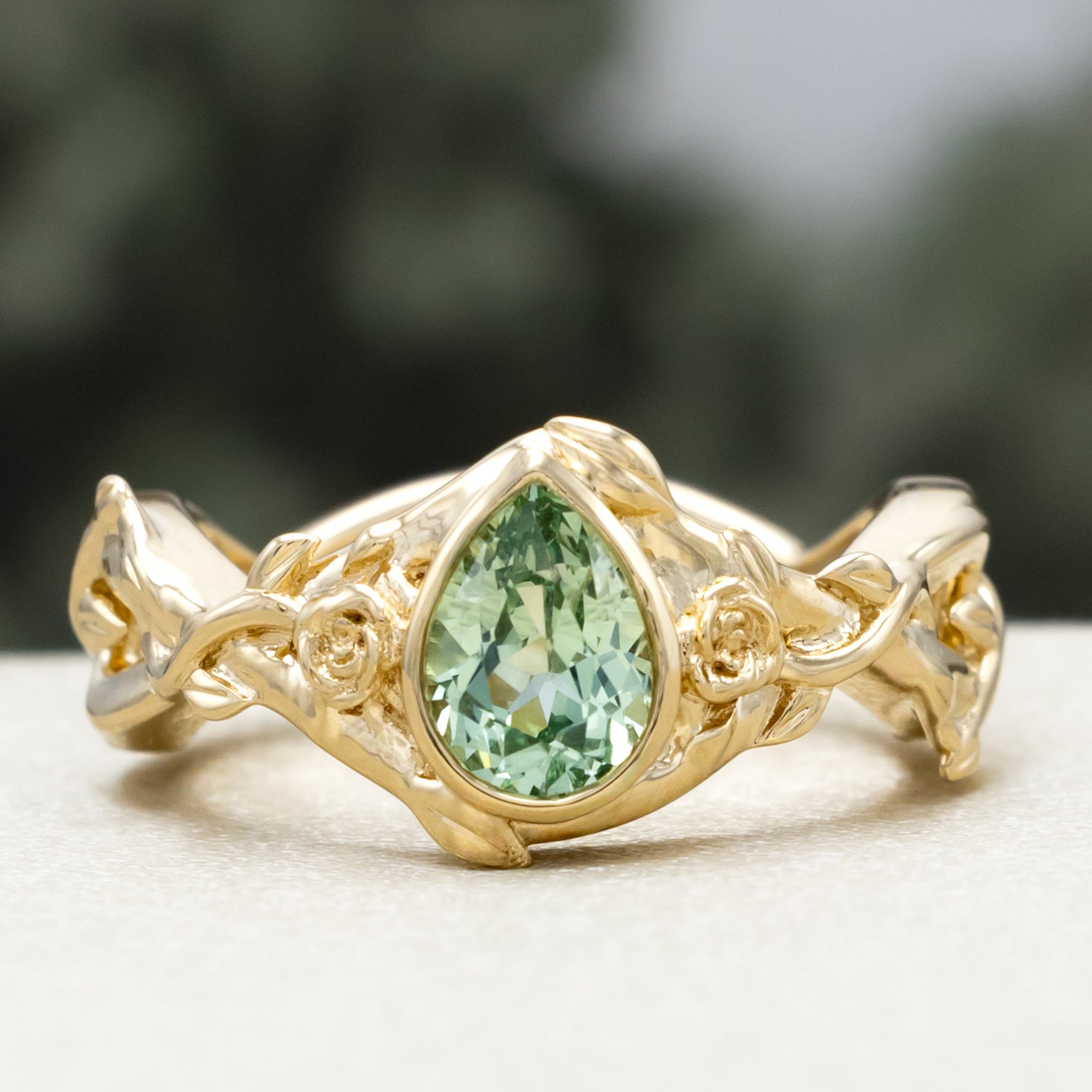 Gold ring with pear lab sapphire and rose/vine accents on a blurred background
