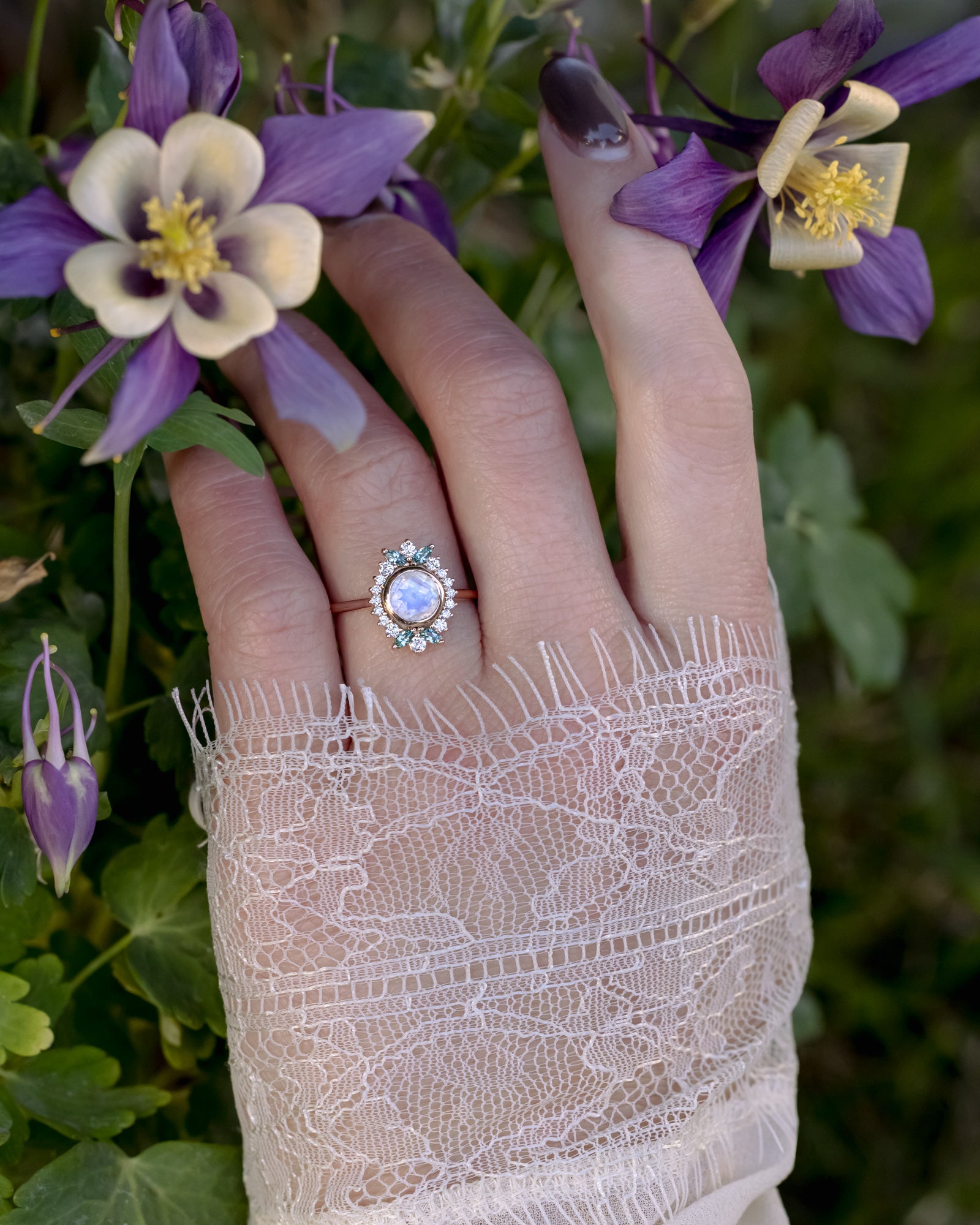 Hand wearing a rose gold ring with a moonstone, surrounded by purple and white flowers.