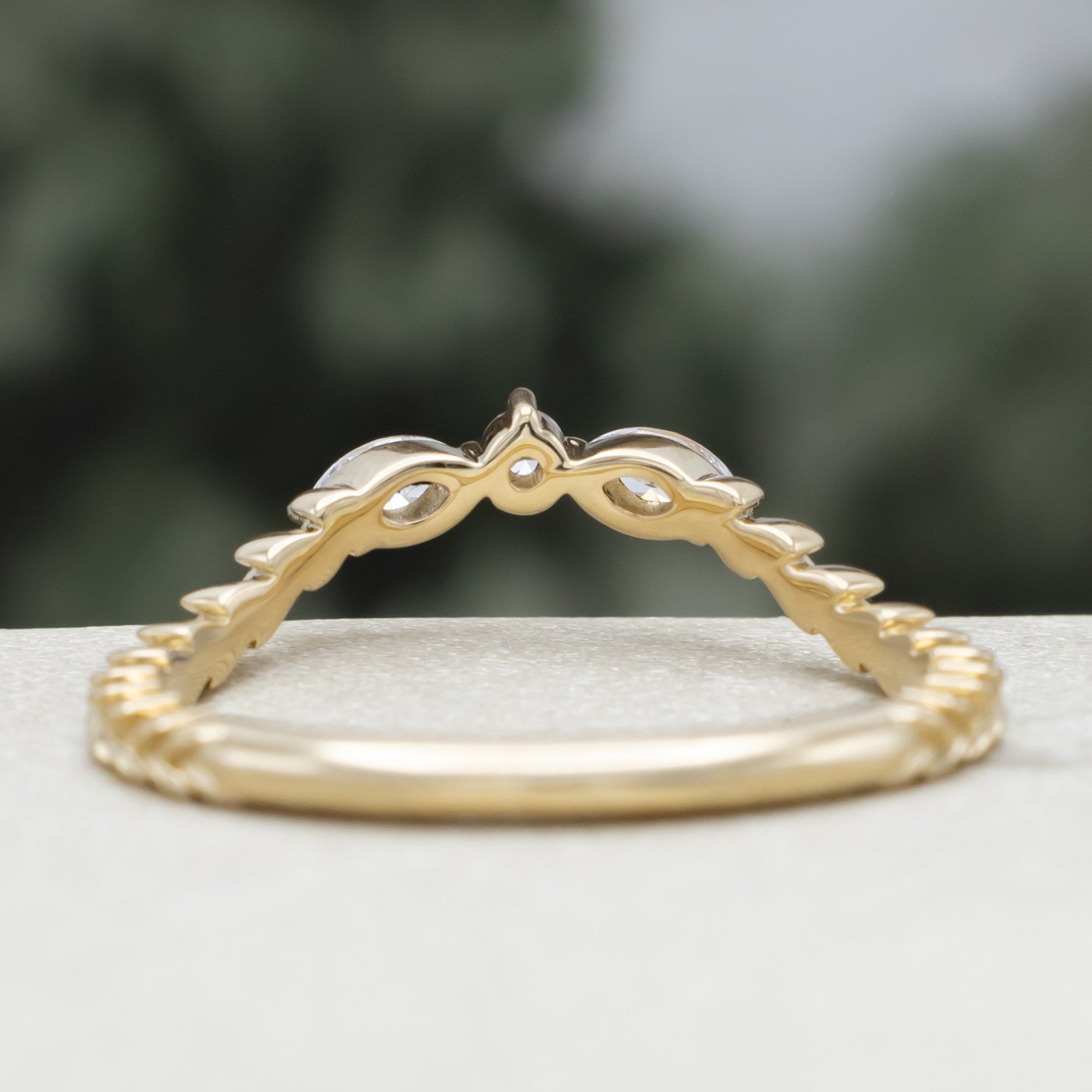 A yellow gold ring tracer with three diamond accents on a vine style band against a blurred green background