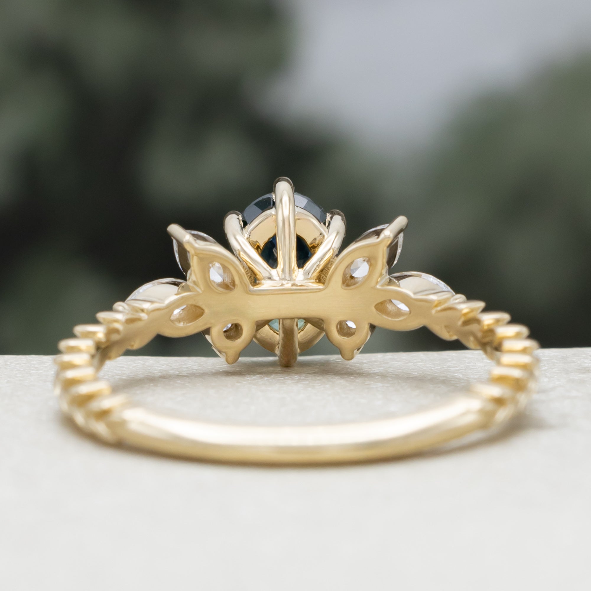A yellow gold ring featuring an oval-cut Montana sapphire with diamond accents on a vine band against a blurred green background.