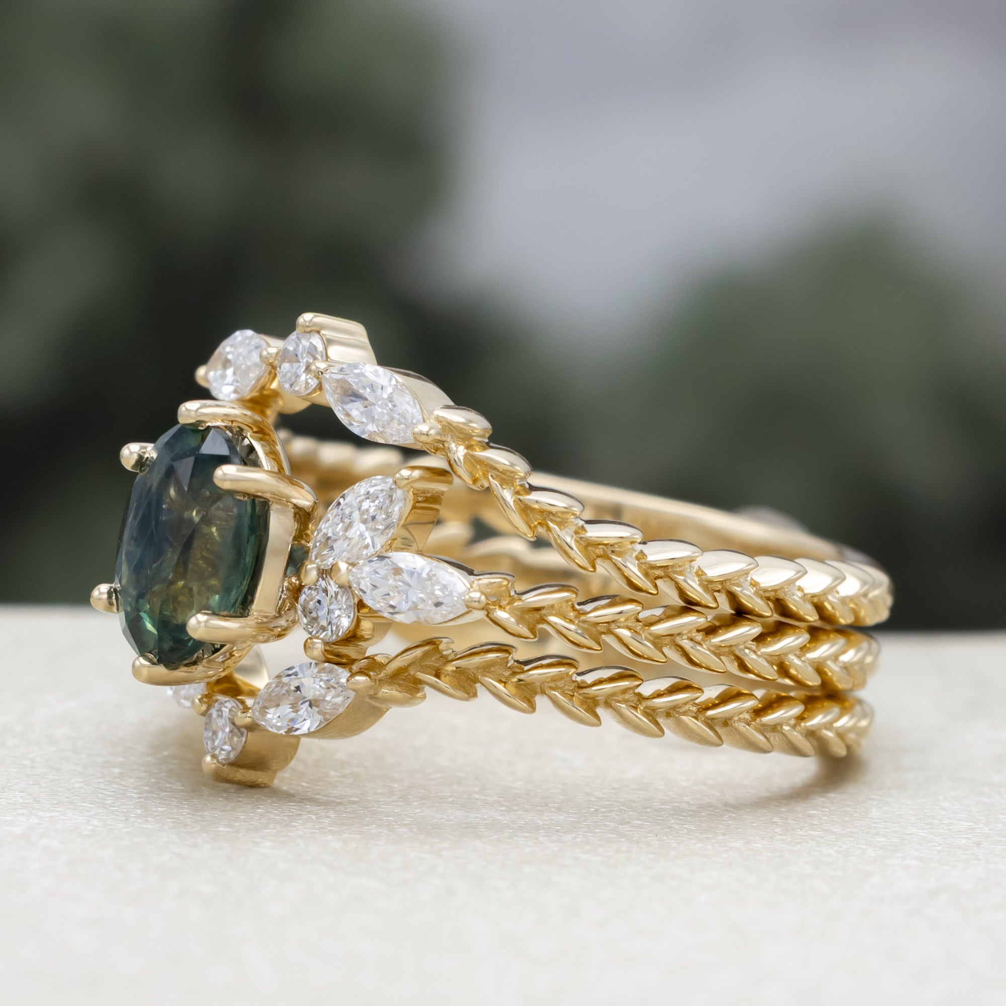 A yellow gold ring featuring an oval-cut Montana sapphire with diamond accents and tracer on a vine band against a blurred green background.