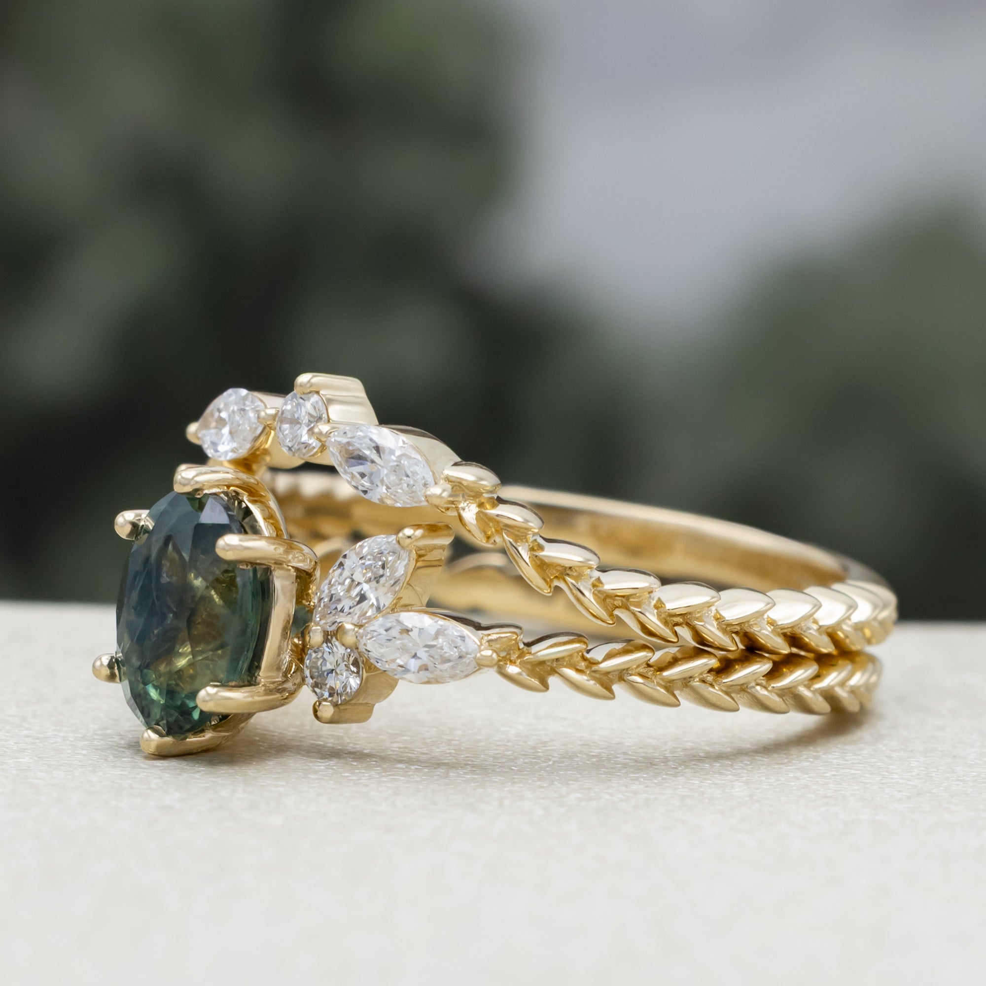 A yellow gold ring featuring an oval-cut Montana sapphire with diamond accents and tracer on a vine band against a blurred green background.