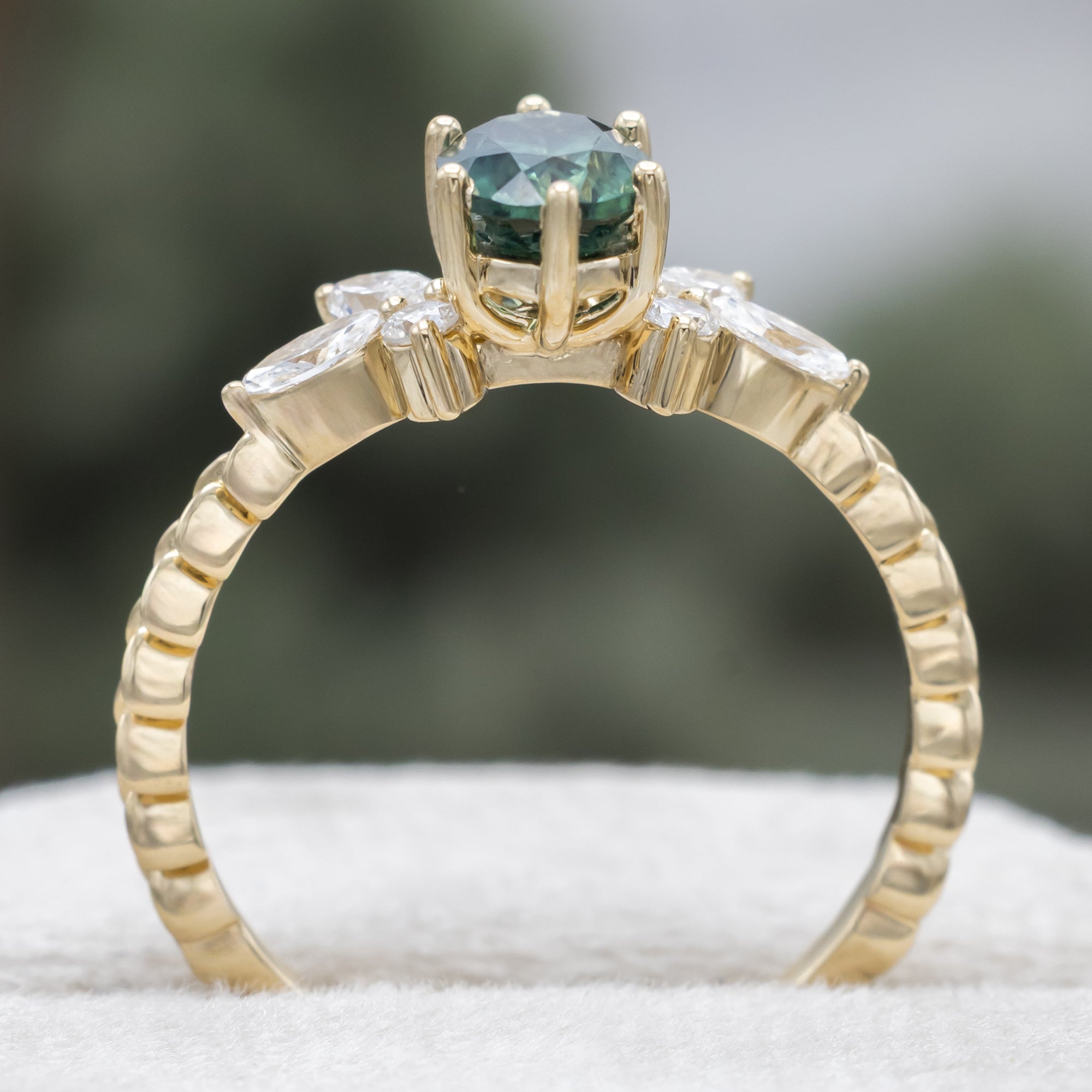 A yellow gold ring featuring an oval-cut Montana sapphire with diamond accents on a vine band against a blurred green background.