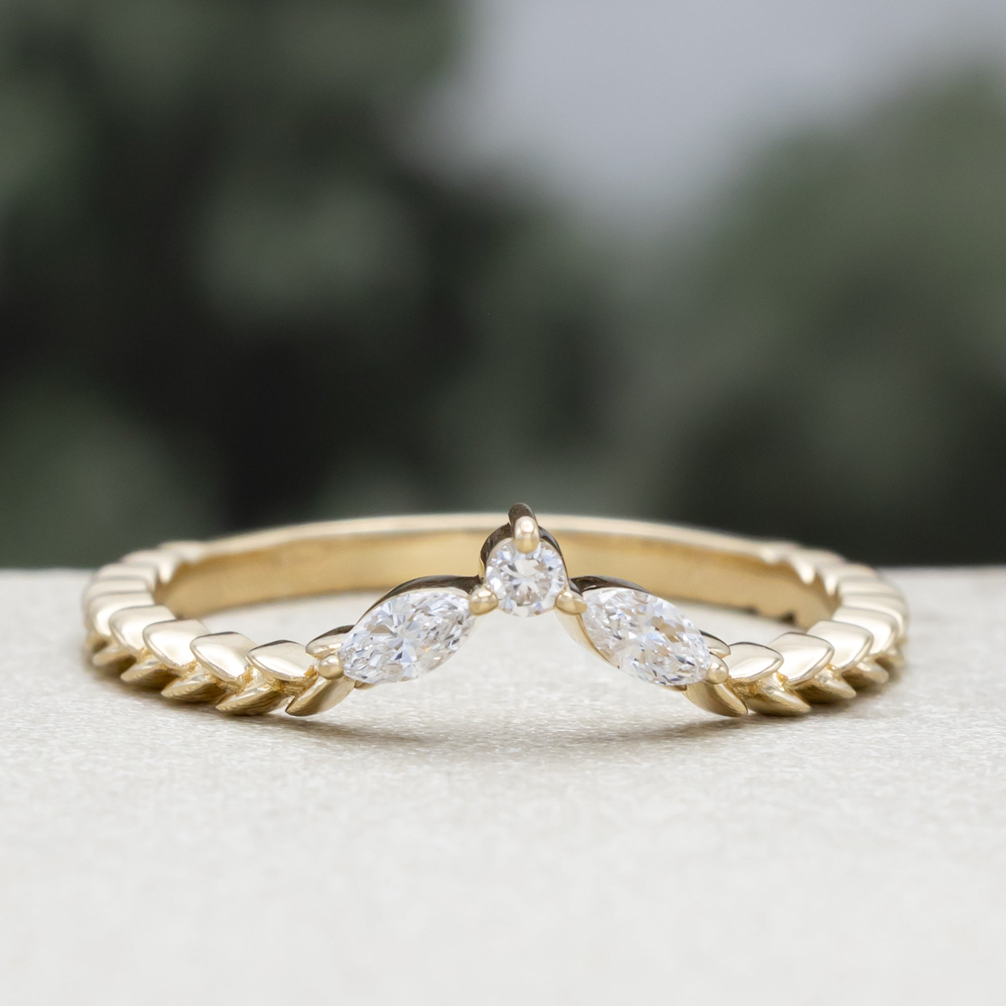 A yellow gold ring tracer with three diamond accents on a vine style band against a blurred green background
