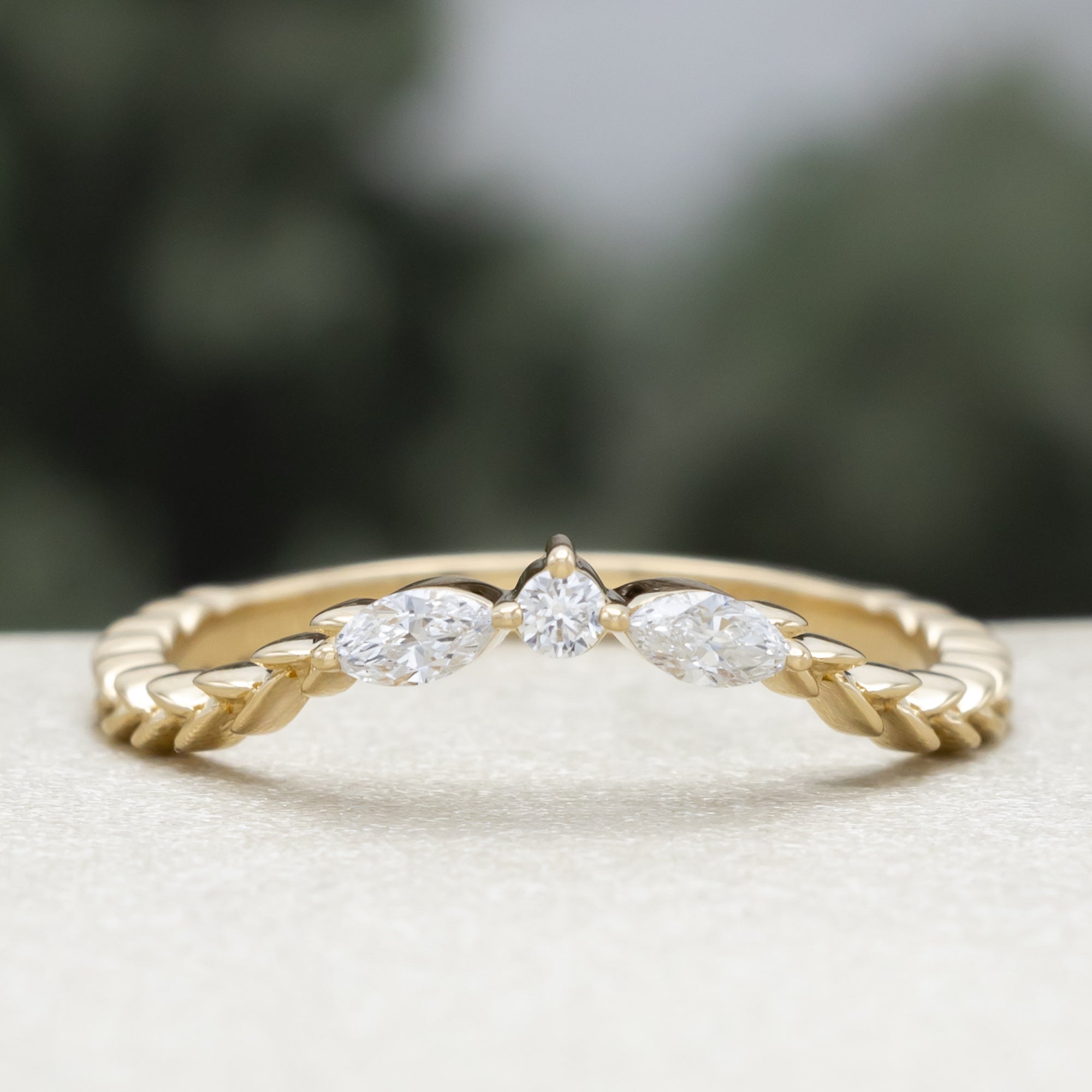 A yellow gold ring tracer with three diamond accents on a vine style band against a blurred green background