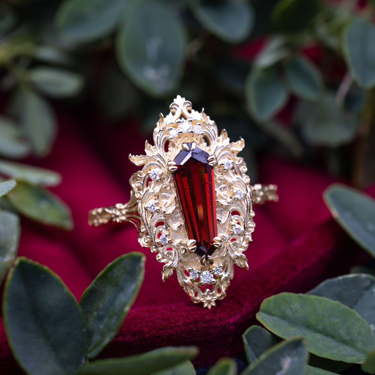 A gold ring with a garnet center stone sitting on a red cushion against a foliage background