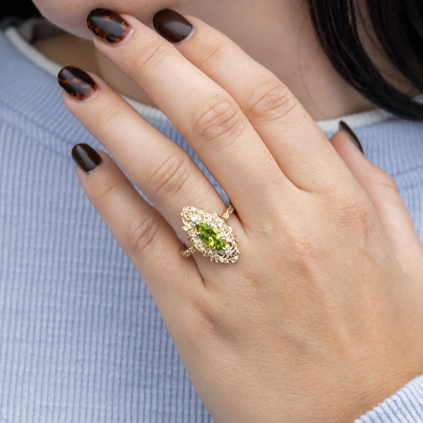 Close-up of a hand wearing an ornate ring with a green peridot, against a light blue background.