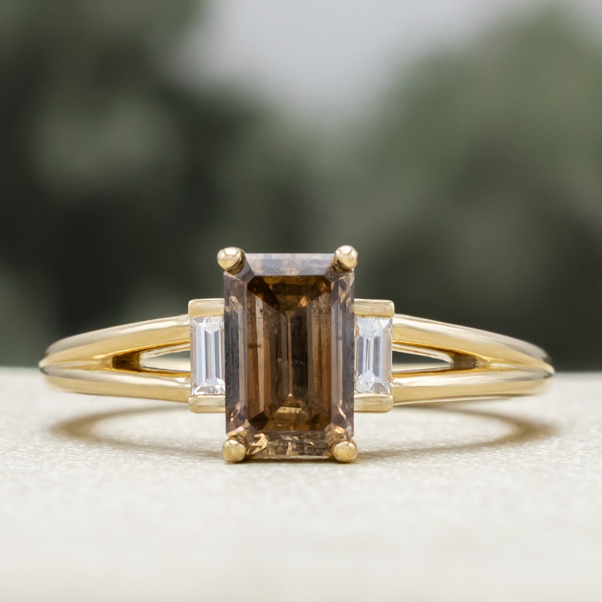 Gold ring with champagne diamond centerstone & diamond accent against a blurred green background