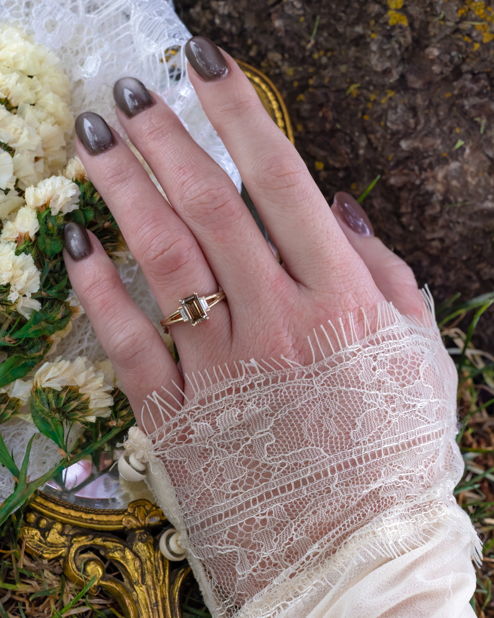 Hand wearing a gold champagne diamond ring with lace sleeve and flowers in the background